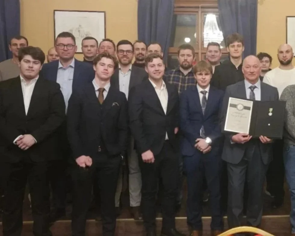 En gruppe menn i formelle klær, noen i dress, samles i et rom med gule vegger og bilder på vegg, en av dem holder en diplom i hånden, en pris Langset fikk for inkludering og mangfold.