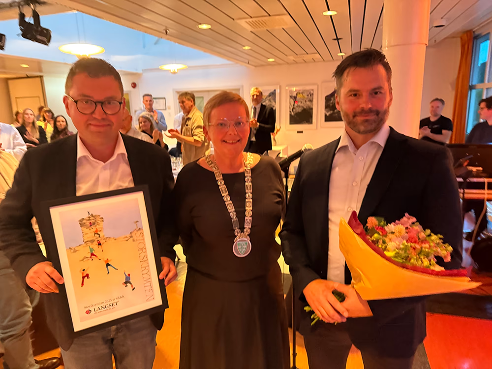 I 2025 fikk vi næringsprisen Storskrymten – en anerkjennelse til alle ansatte i Langset Mek AS. Bildet viser to ansatte fra bedriften som mottar prisen, og som holder diplom og blomster i hendene, sammen med prisutdeler.