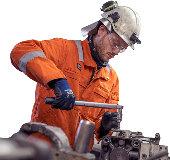 En mann i orange arbeidsdress og hjelm arbeider med et metallstykke i en verkstedsetting.