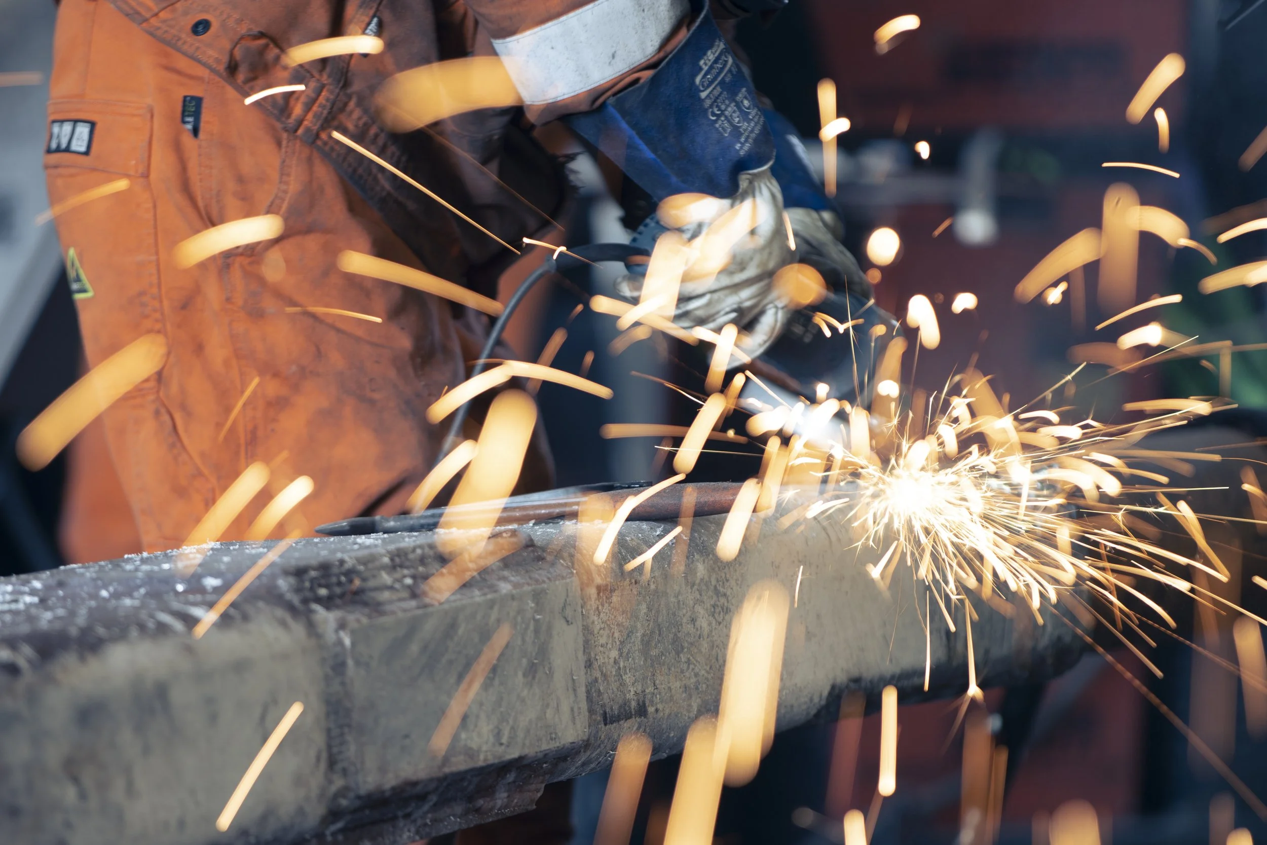 En person i oransje arbeidsdress som sveiser en metallbjelke. Det spruter ut gnister.