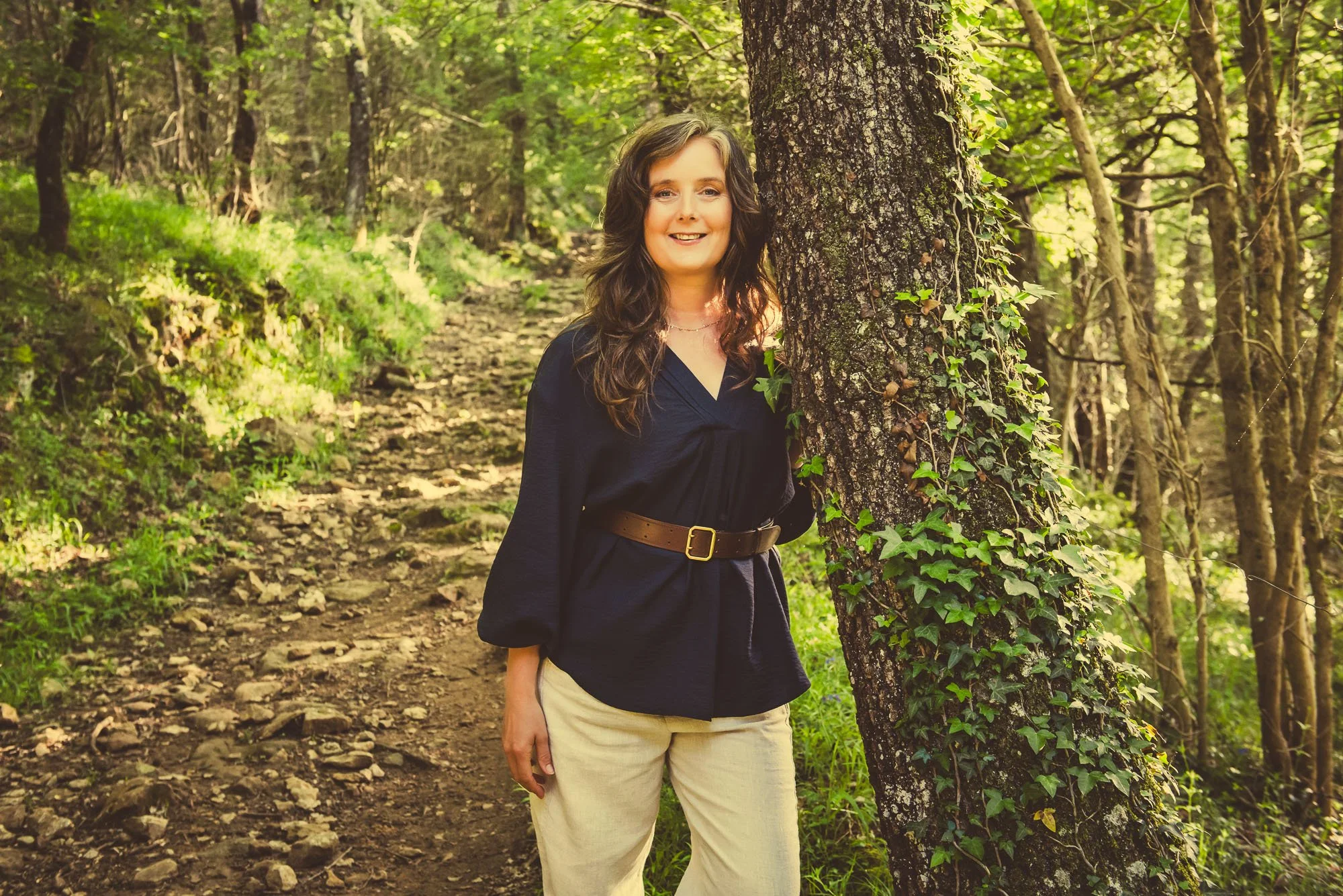 A woman smiling standing outdoors behind a tree with green leaves in a wooded area.