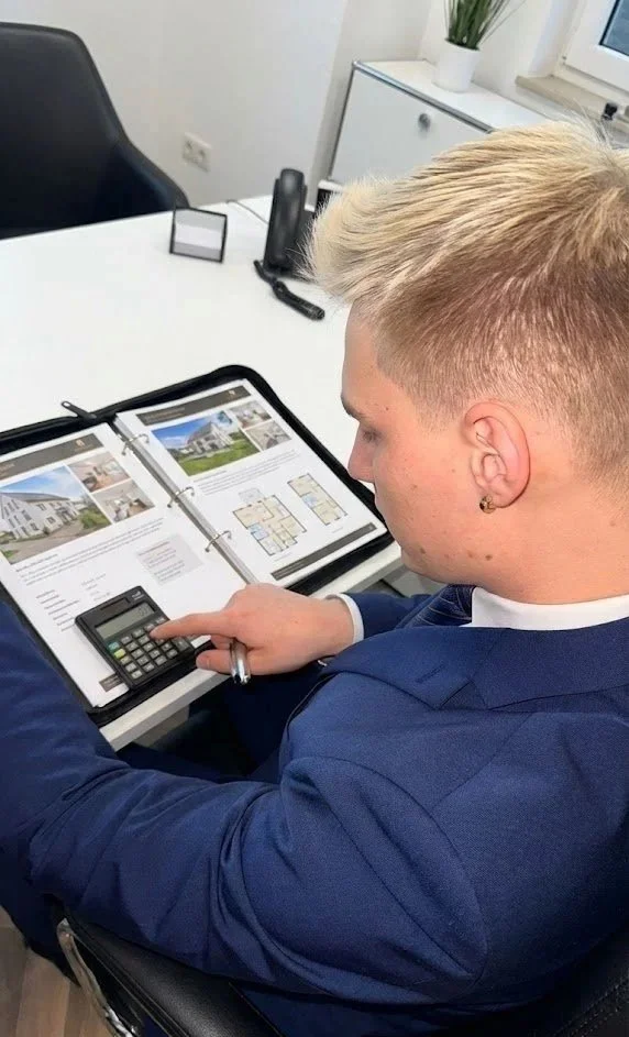Junge in Anzug sitzt an Schreibtisch, nutzt Taschenrechner und liest Immobilienplan, im Büro.