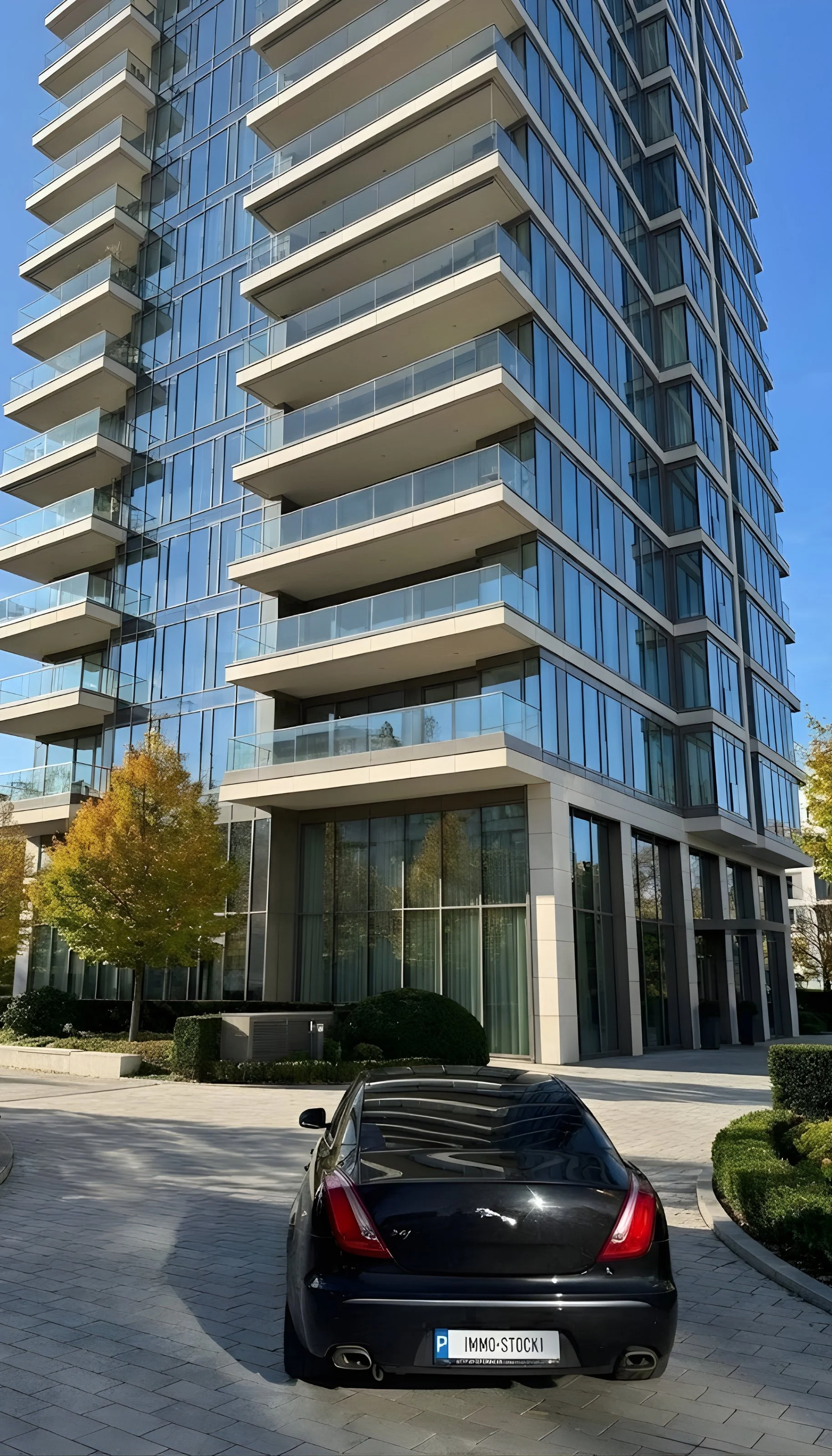 Hochhaus mit mehreren Balkonen und großen Glasfenstern, davor ein schwarzer Sportwagen auf gepflastertem Parkplatz, umgeben von Büschen und Bäumen, bei sonnigem Wetter.