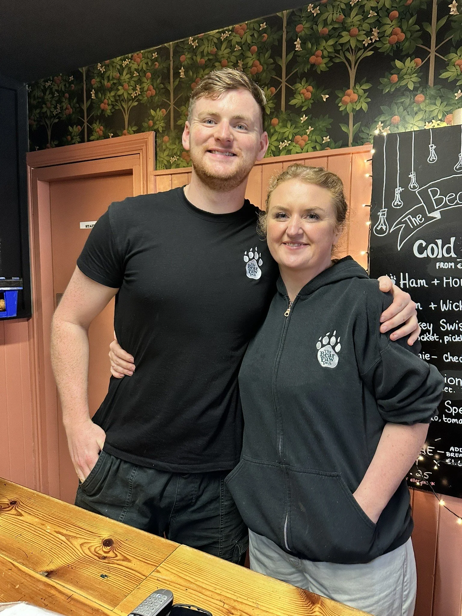 A man and woman standing inside a café or restaurant, smiling and posing for the photo. They are wearing black clothing with a paw print logo that says "The Bear Paw". The background includes a blackboard menu and decorative wallpaper with a floral p