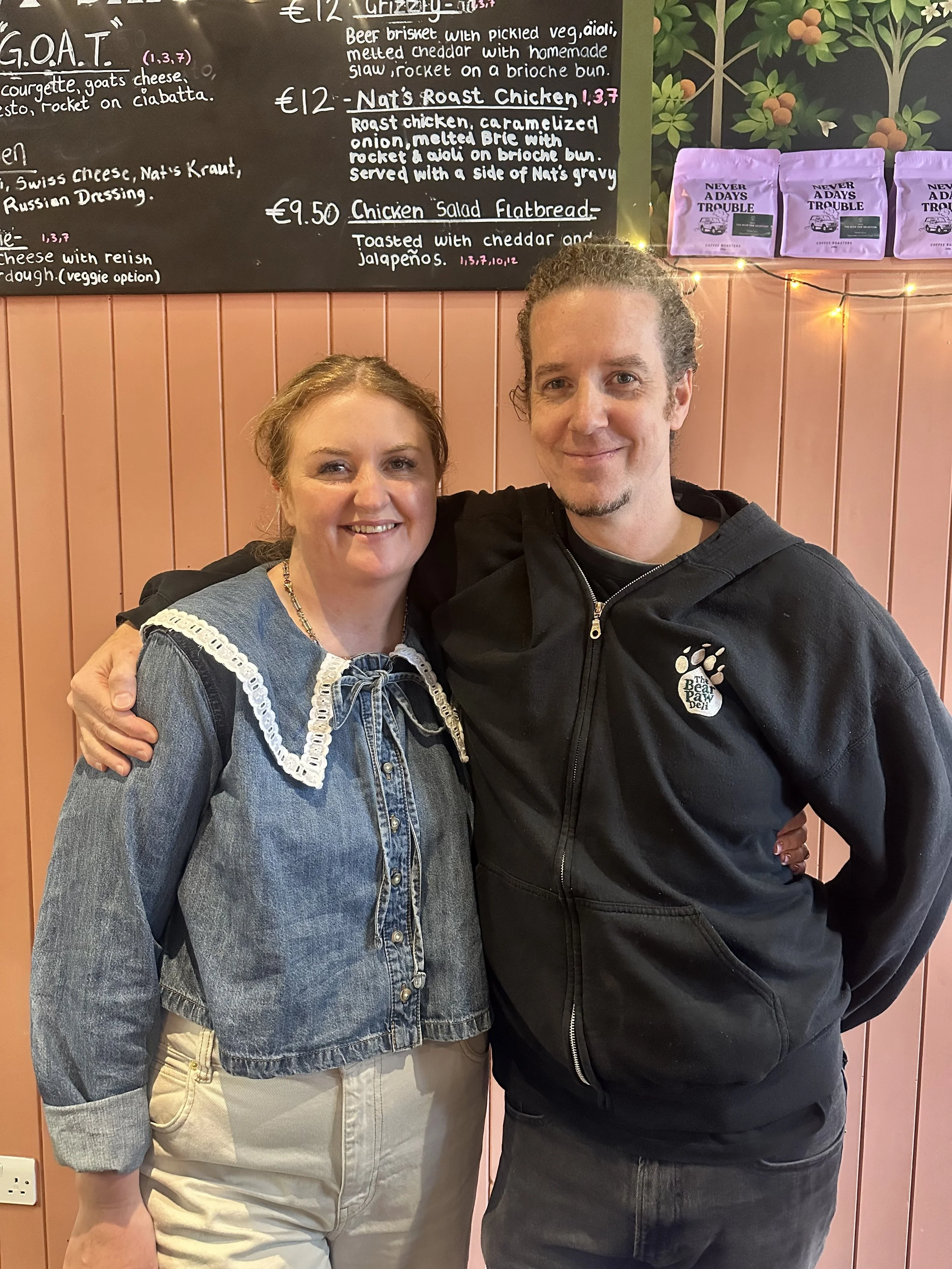 A woman and a man standing together in a cozy cafe, smiling at the camera. The woman has light brown hair, wearing a denim jacket with a lace collar and beige pants. The man has curly hair tied back, wearing a black hoodie with a paw print logo. Behi