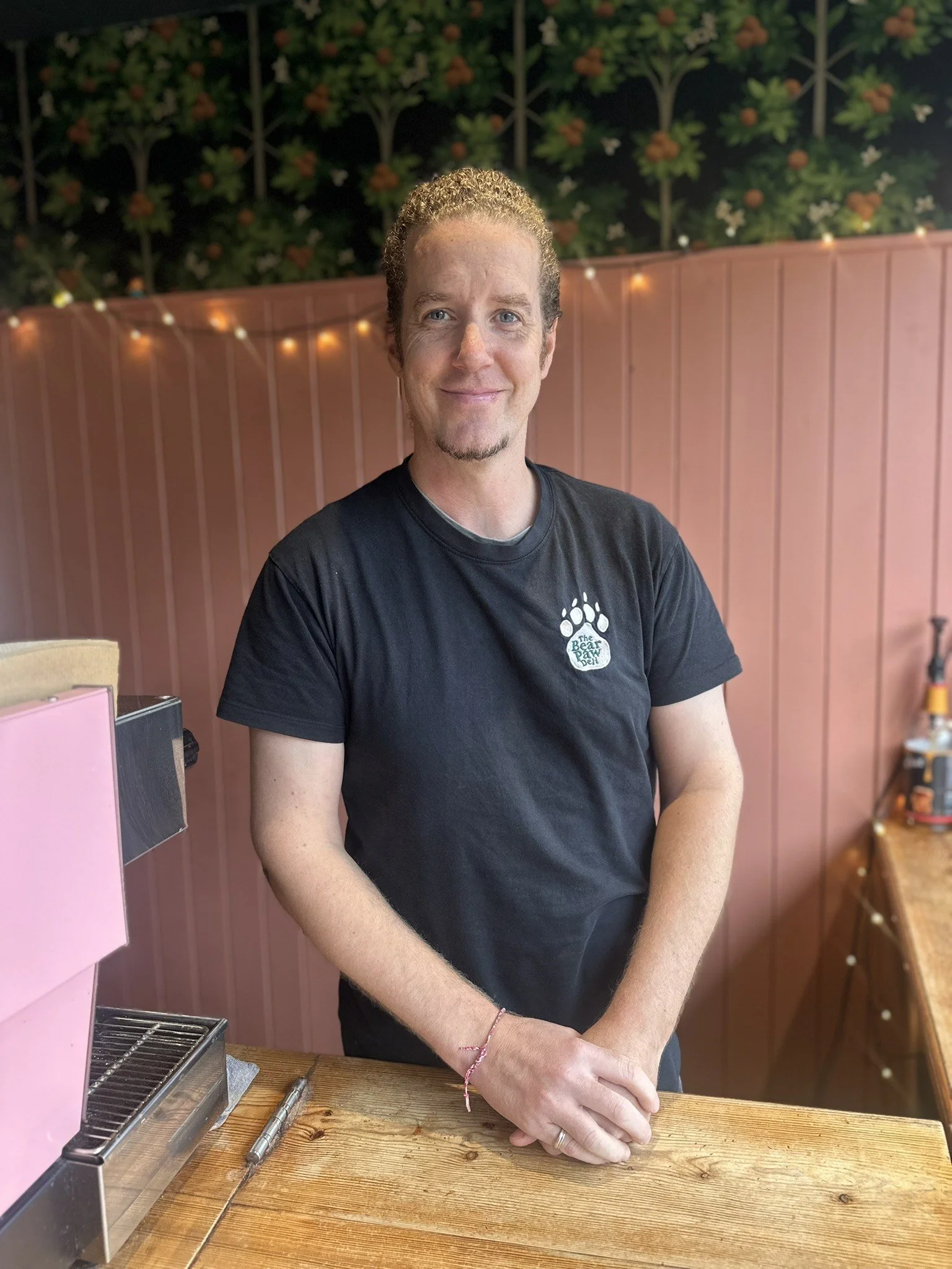 A man with curly light brown hair and a goatee, smiling, standing behind a wooden counter with a pink appliance and a metal tool, against a pink wall with string lights and decorative greenery.