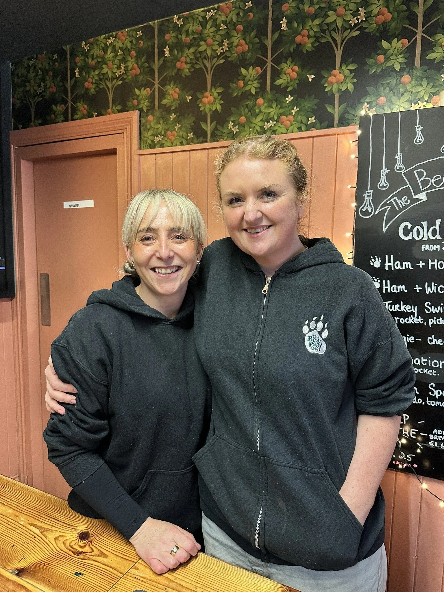 Two women in black hoodies smiling and hugging in a cozy interior with a wooden counter and a blackboard menu behind them.