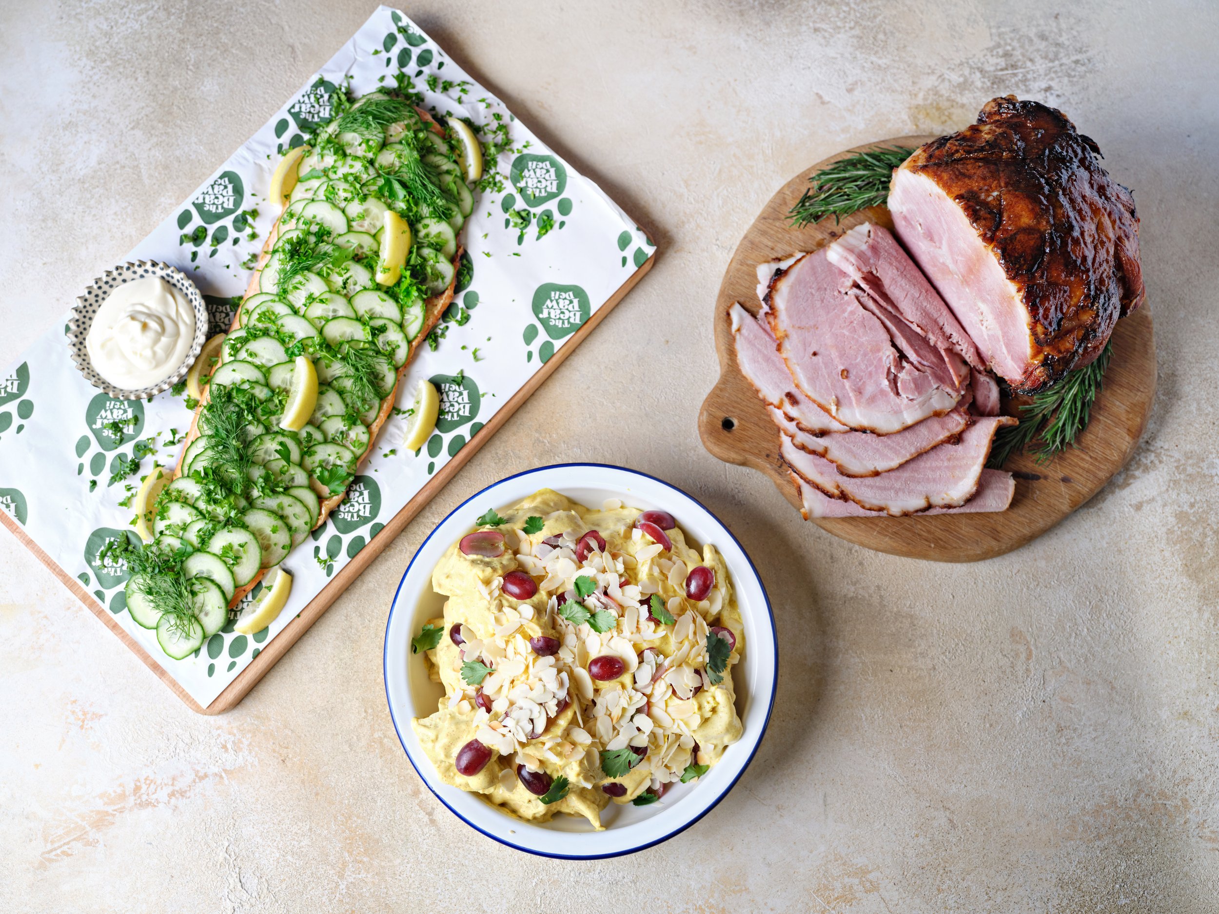 A platter of cucumber and dill open-faced sandwich, a bowl of egg salad with almonds and pomegranate seeds, and a roasted ham on a wooden cutting board with rosemary garnish.