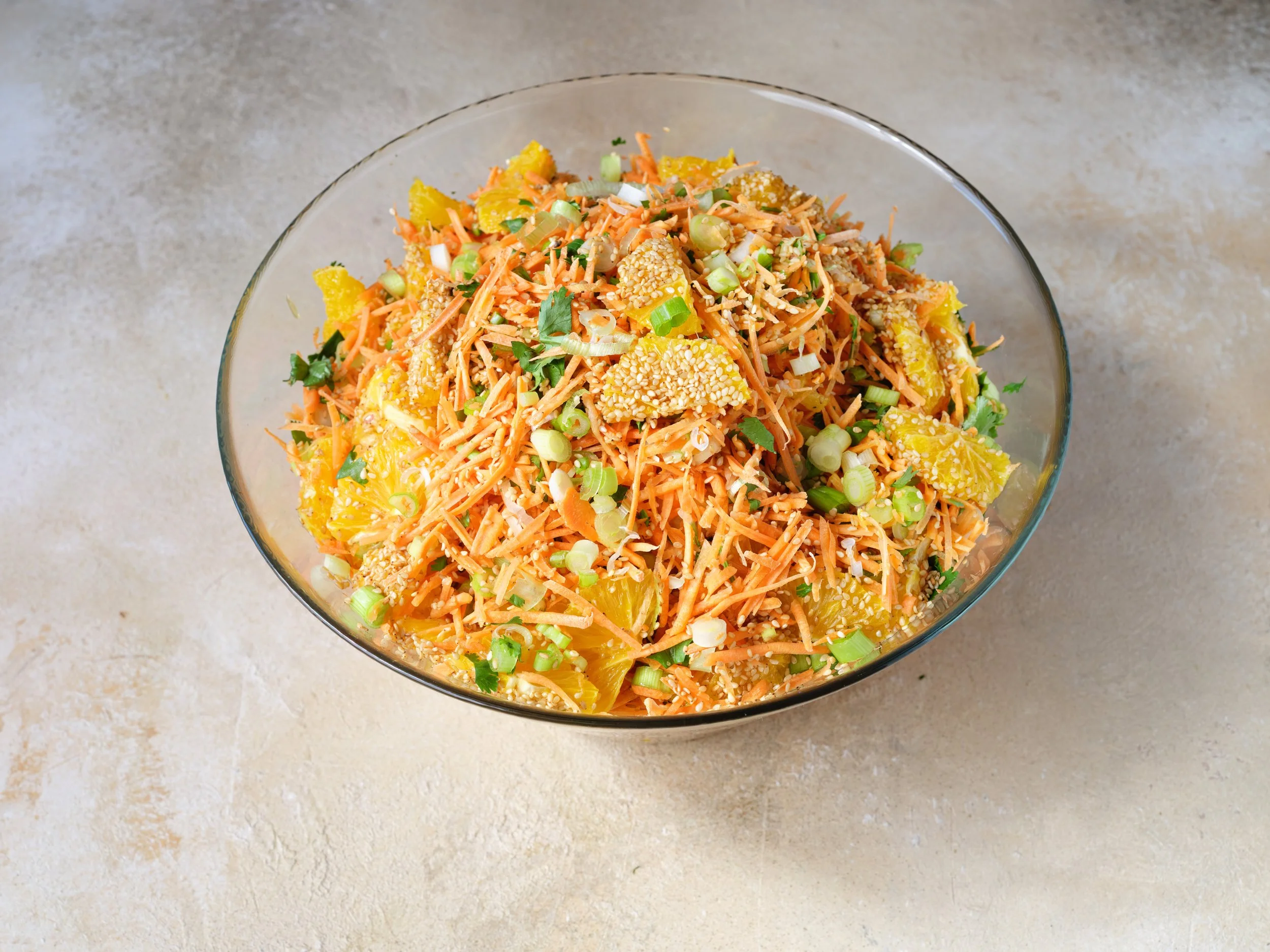 A glass bowl filled with shredded carrots, chopped green onions, chopped cilantro, sesame seeds, and yellow pickled vegetables.