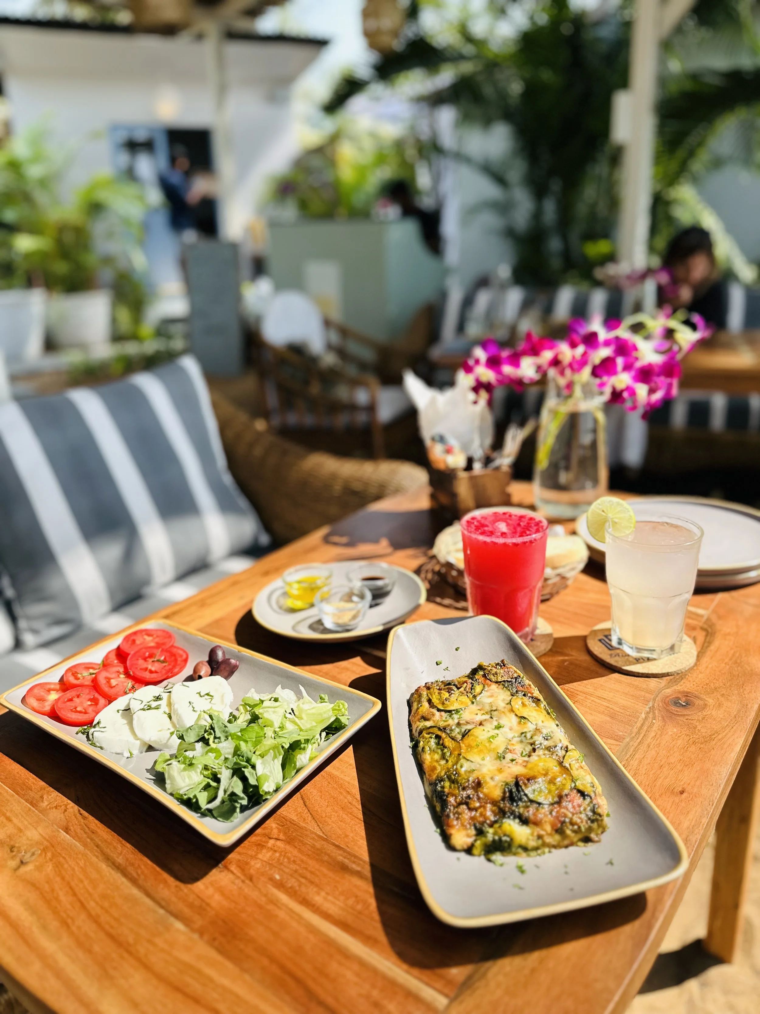 A table with a salad, baked dish, and drinks at an outdoor cafe or restaurant, with flowers and greenery in the background.