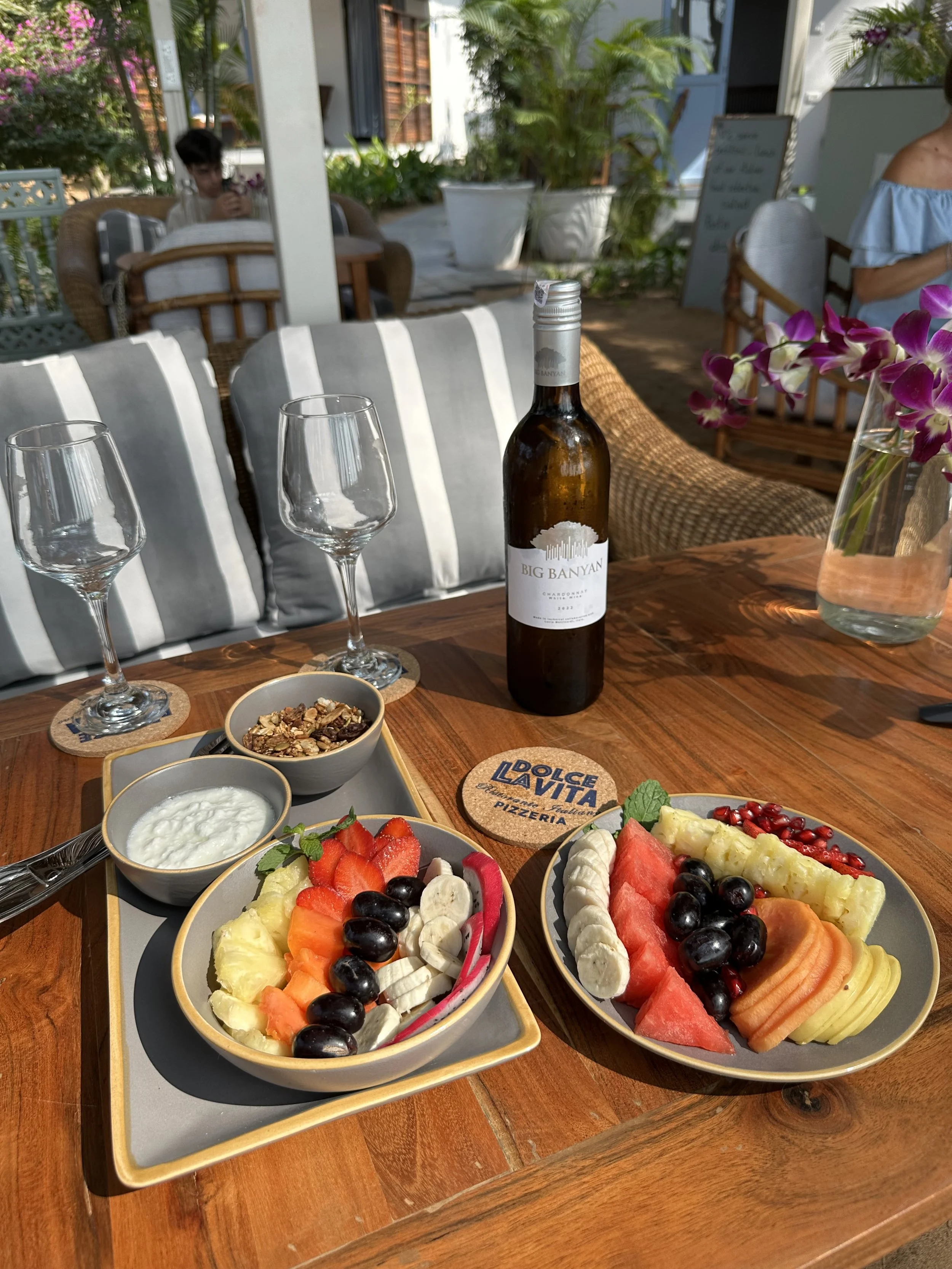 Two plates of fresh sliced fruit including watermelon, banana, grapes, pineapple, honeydew, cantaloupe, dragon fruit, and pomegranate seeds on a wooden table. A bottle of white wine and two empty wine glasses are also on the table. In the background, a person is sitting and looking at their phone, with plants and outdoor seating visible.