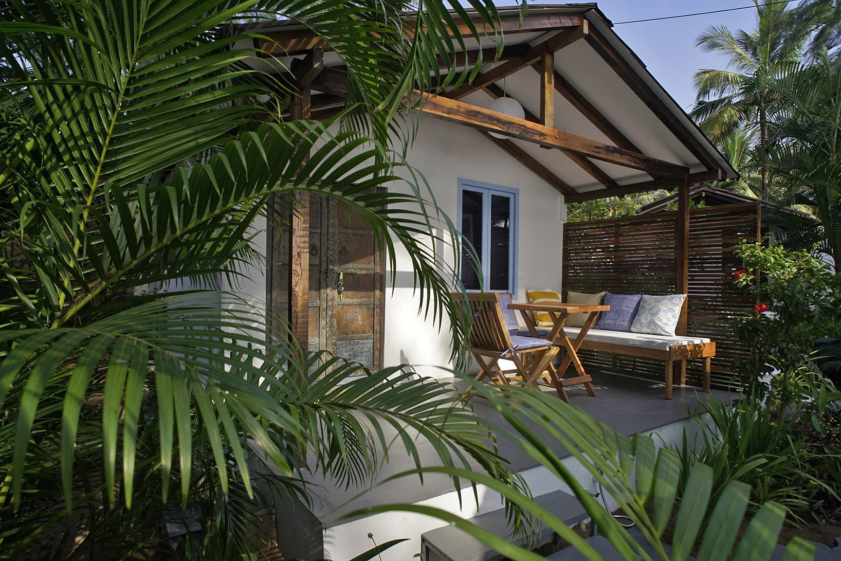 A cozy outdoor patio of a small house surrounded by lush green tropical plants. The patio has a wooden bench with cushions, a small wooden table, and a wooden chair. The house has white walls, a window, and a rustic wooden door. The roof has exposed wooden beams.