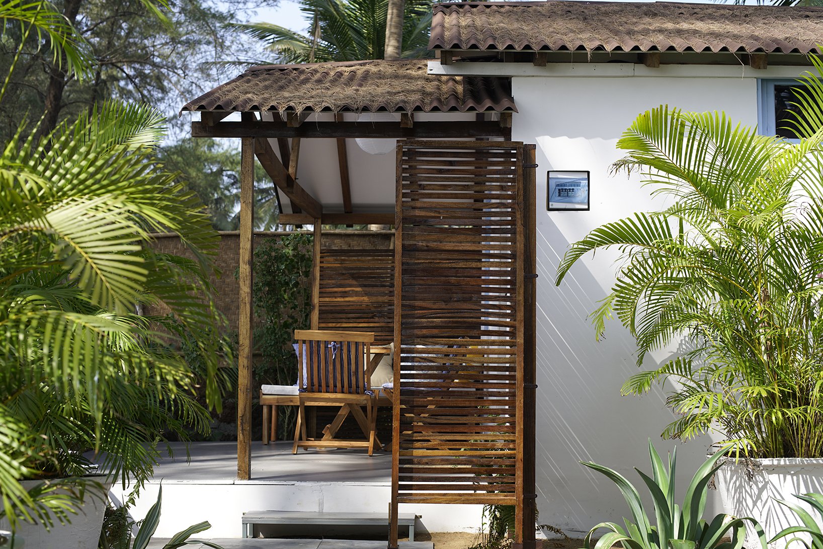 Small patio with wooden chair and slatted wooden privacy screen, surrounded by lush green potted plants, next to a white house wall with a small framed picture.
