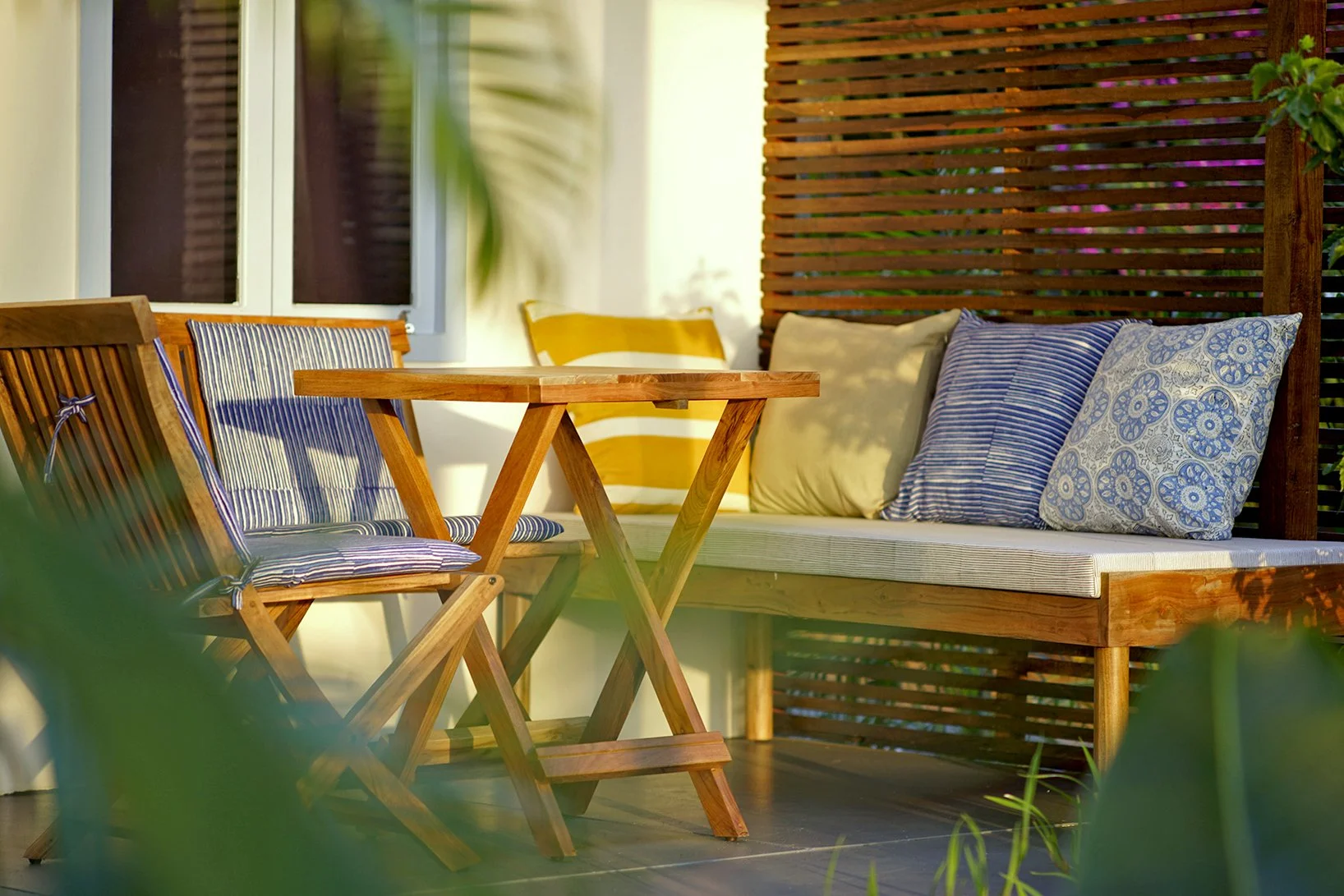 Outdoor patio with wooden table, foldable wooden chairs with striped cushions, and a wooden bench with patterned pillows, set against a wooden slatted privacy screen.
