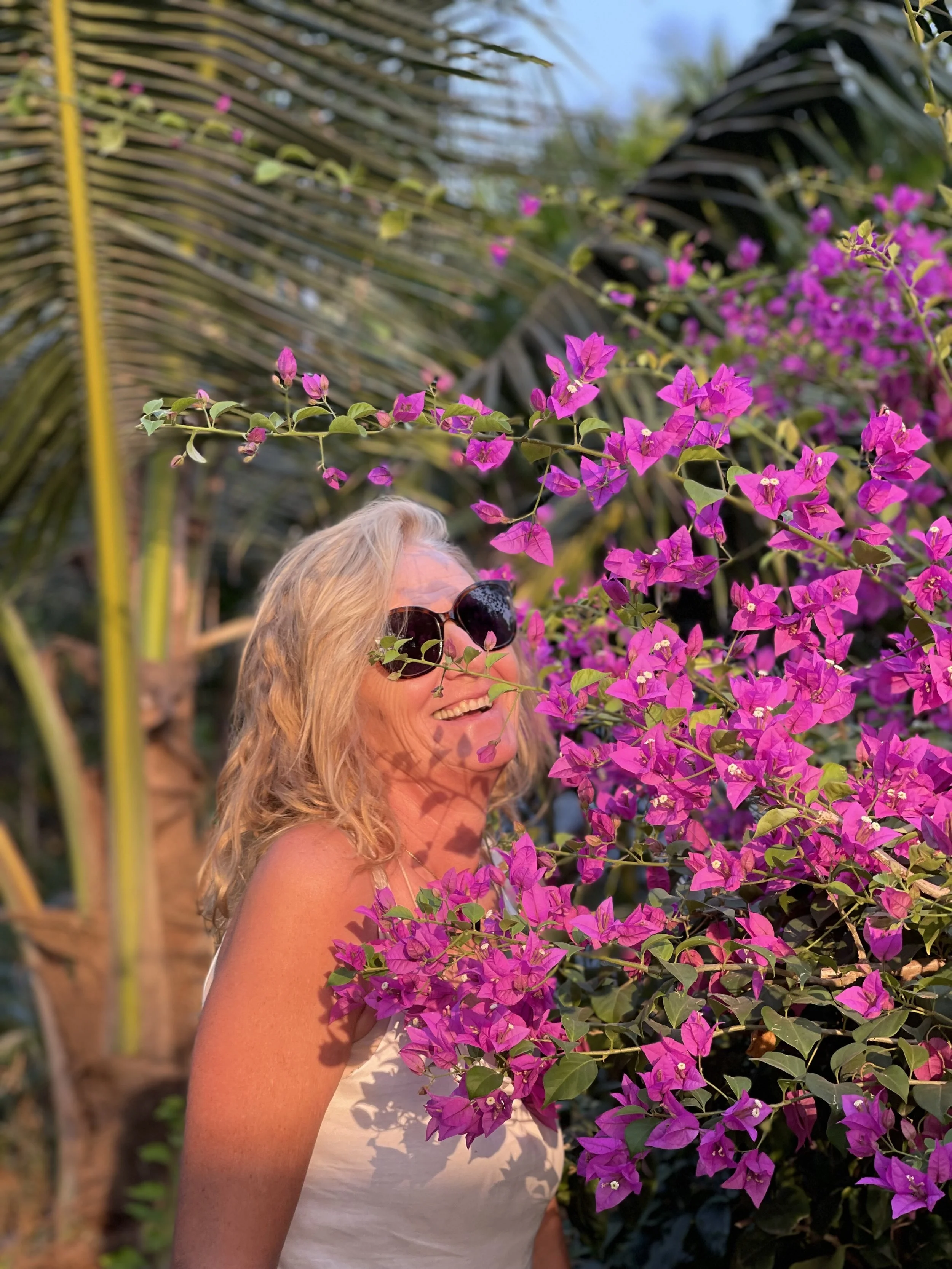 A woman with blonde hair wearing sunglasses smiles among pink bougainvillea flowers and tropical plants.