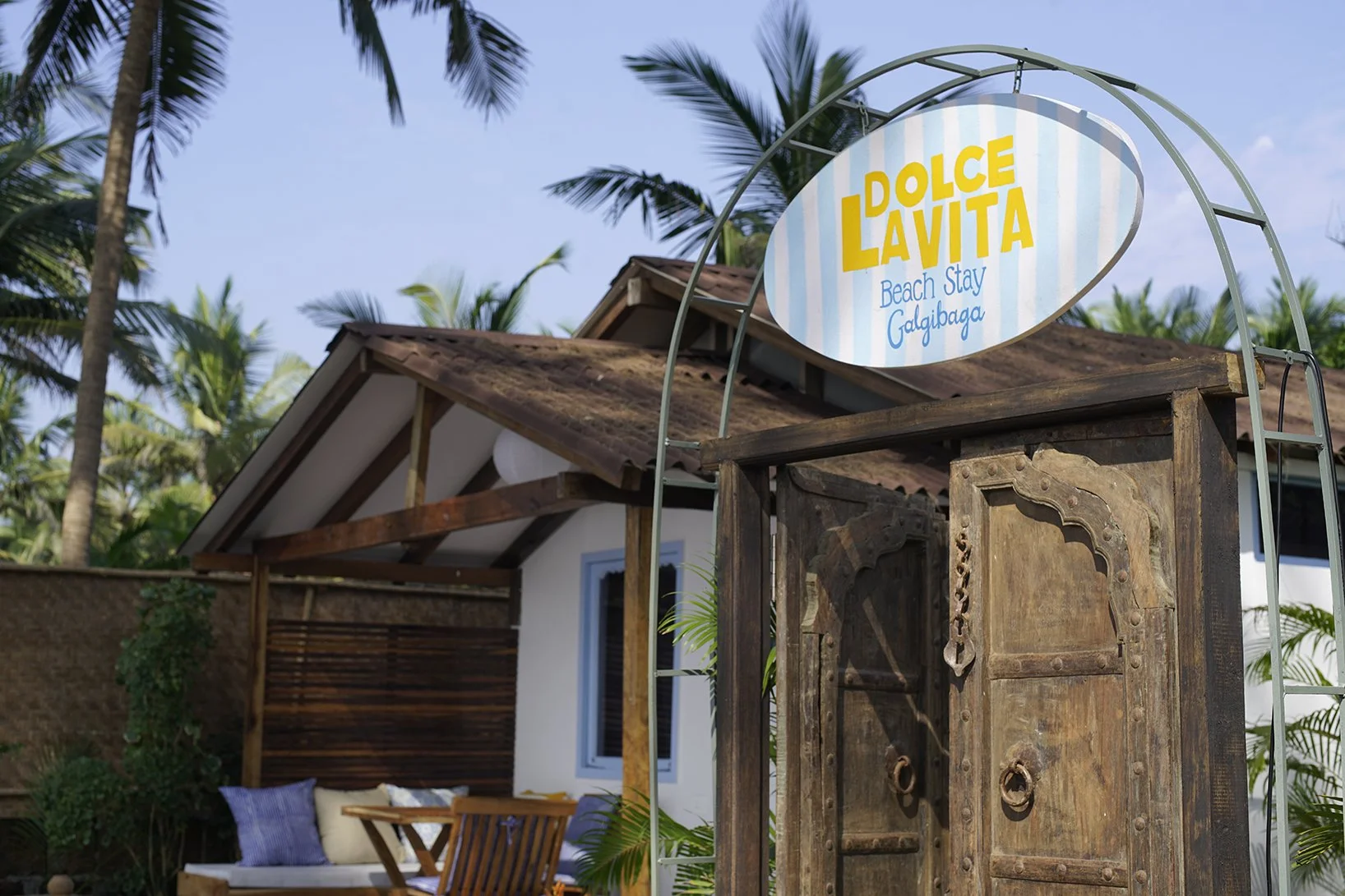 A sign that reads 'DOLCE LAVITA Beach Stay Galibaga' mounted on a frame in front of a small vacation bungalow with a porch, surrounded by palm trees.