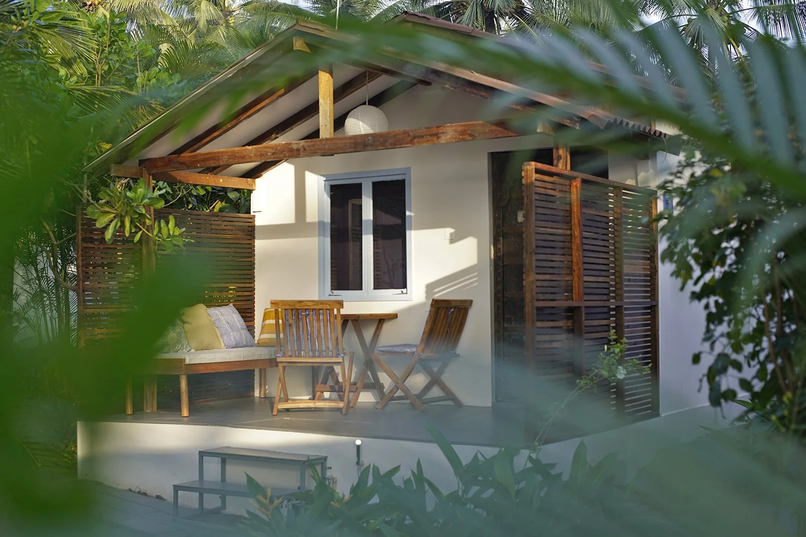 A small wooden house with a porch surrounded by green tropical plants. The porch has a wooden bench with cushions, a small wooden table, and two wooden chairs. The house features a window, a slatted door, and a white exterior wall, with a pendant light hanging from the roof.