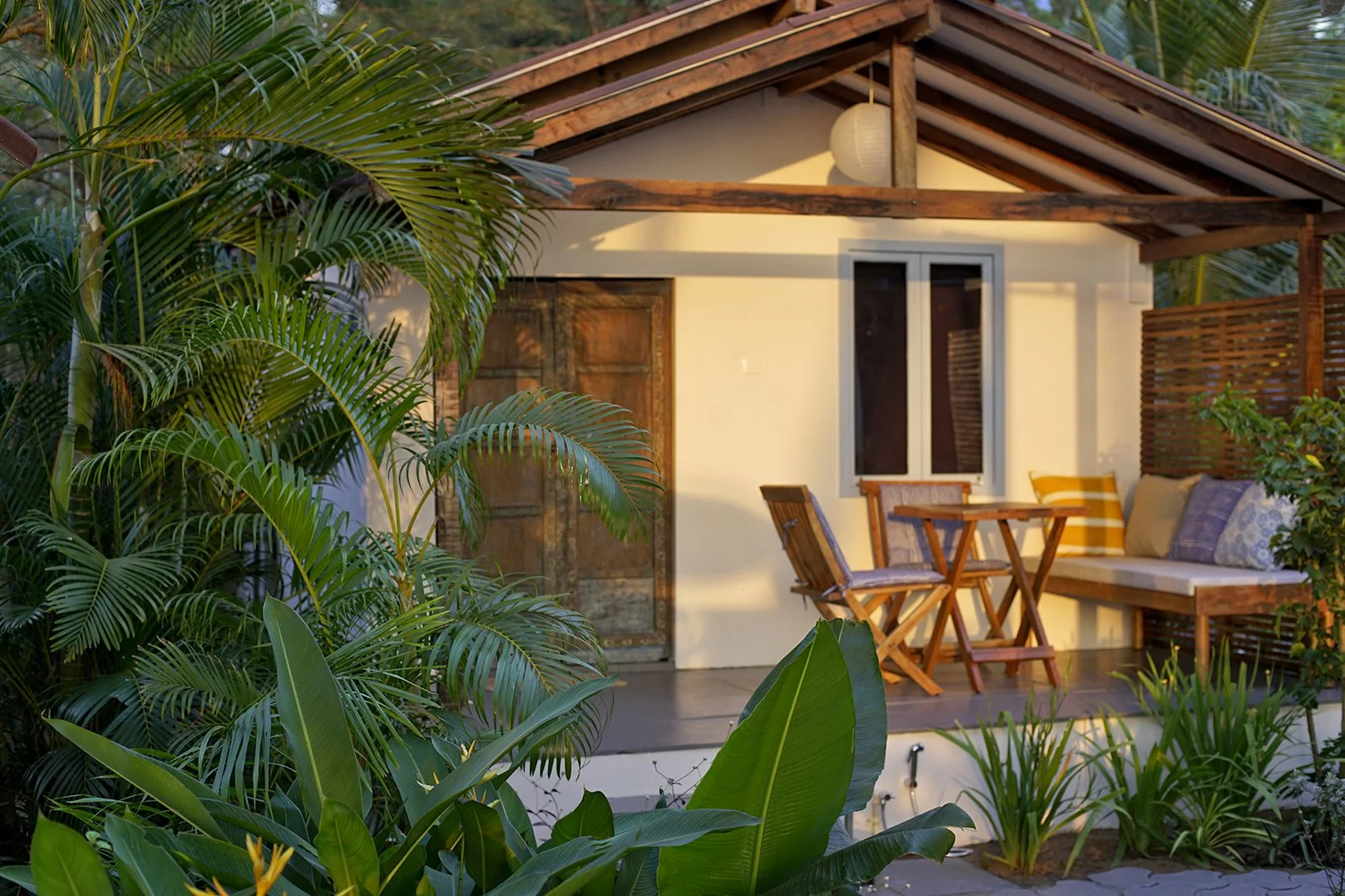 A cozy outdoor patio with wooden furniture and lush green plants surrounding a small house with a white exterior, wooden roof beams, and a window.
