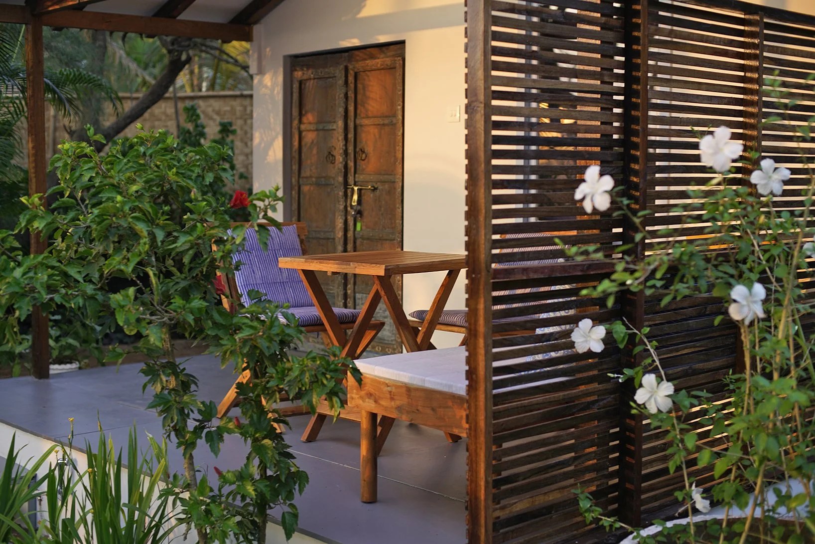 A cozy outdoor patio with wooden furniture, including a small table and chairs with striped cushions, surrounded by lush green plants and white flowers, and a wooden privacy screen. There is a white wall with double rustic wooden doors in the backgro