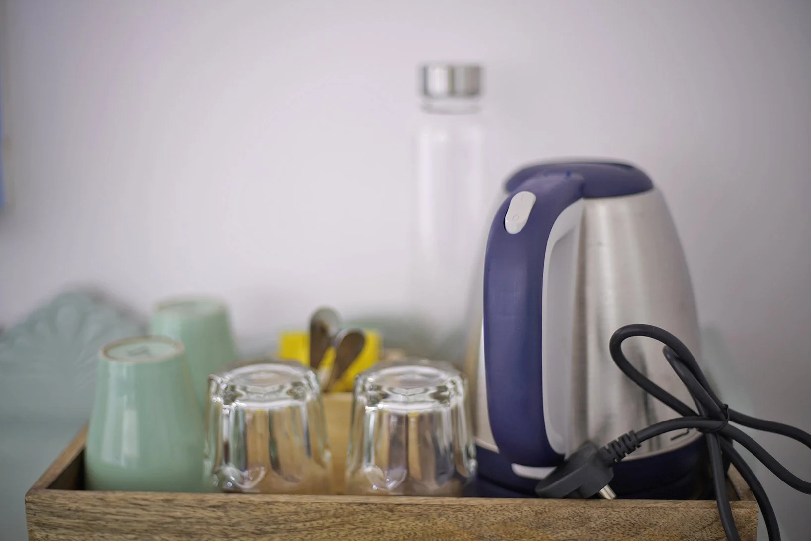 Electric kettle, four glass cups, and mugs inside a wooden tray on a countertop, with a water bottle in the background.