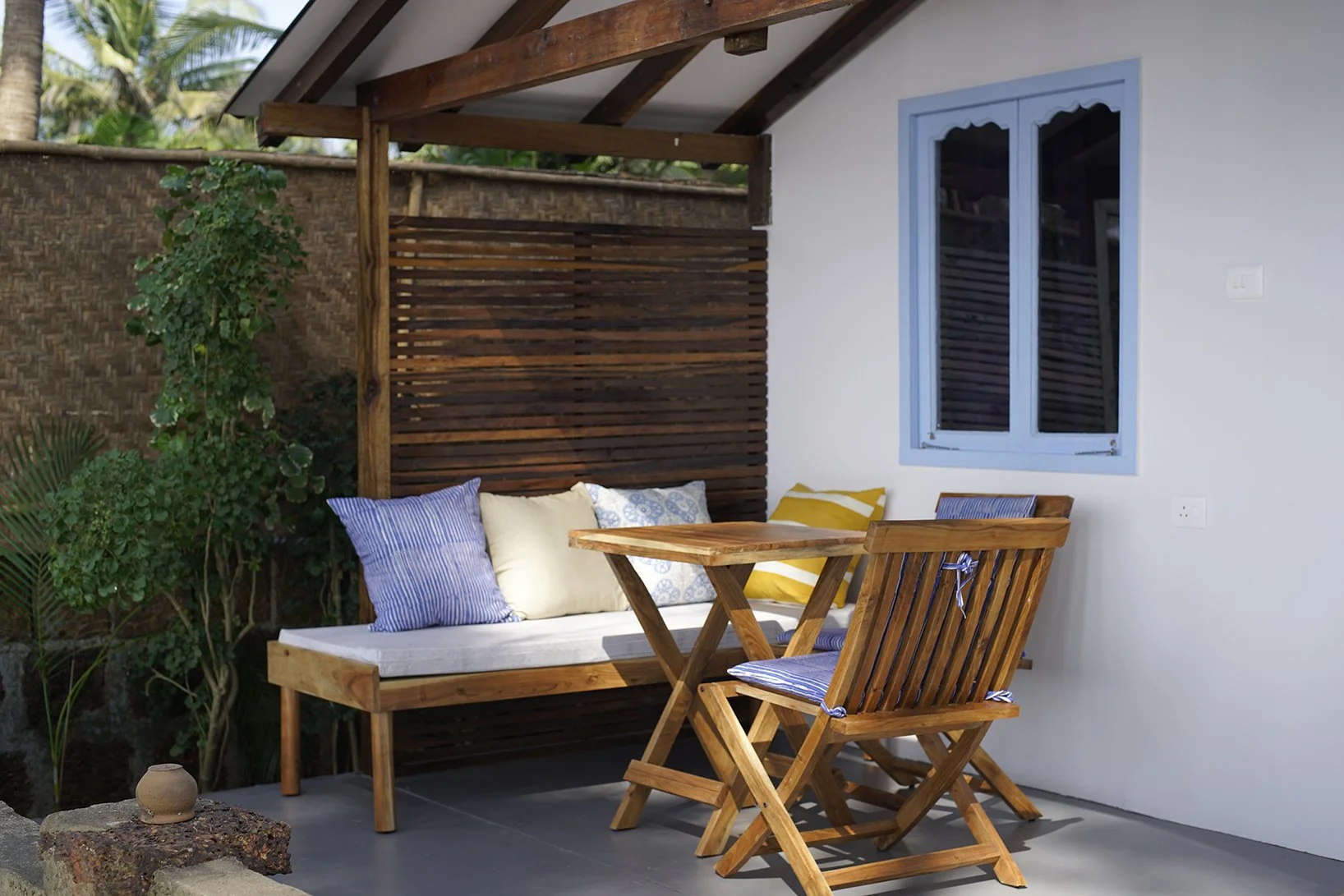 Outdoor patio area with a wooden bench with cushions, a wooden table, and a wooden folding chair in front of a white wall with a blue-framed window, surrounded by greenery including trees and plants.