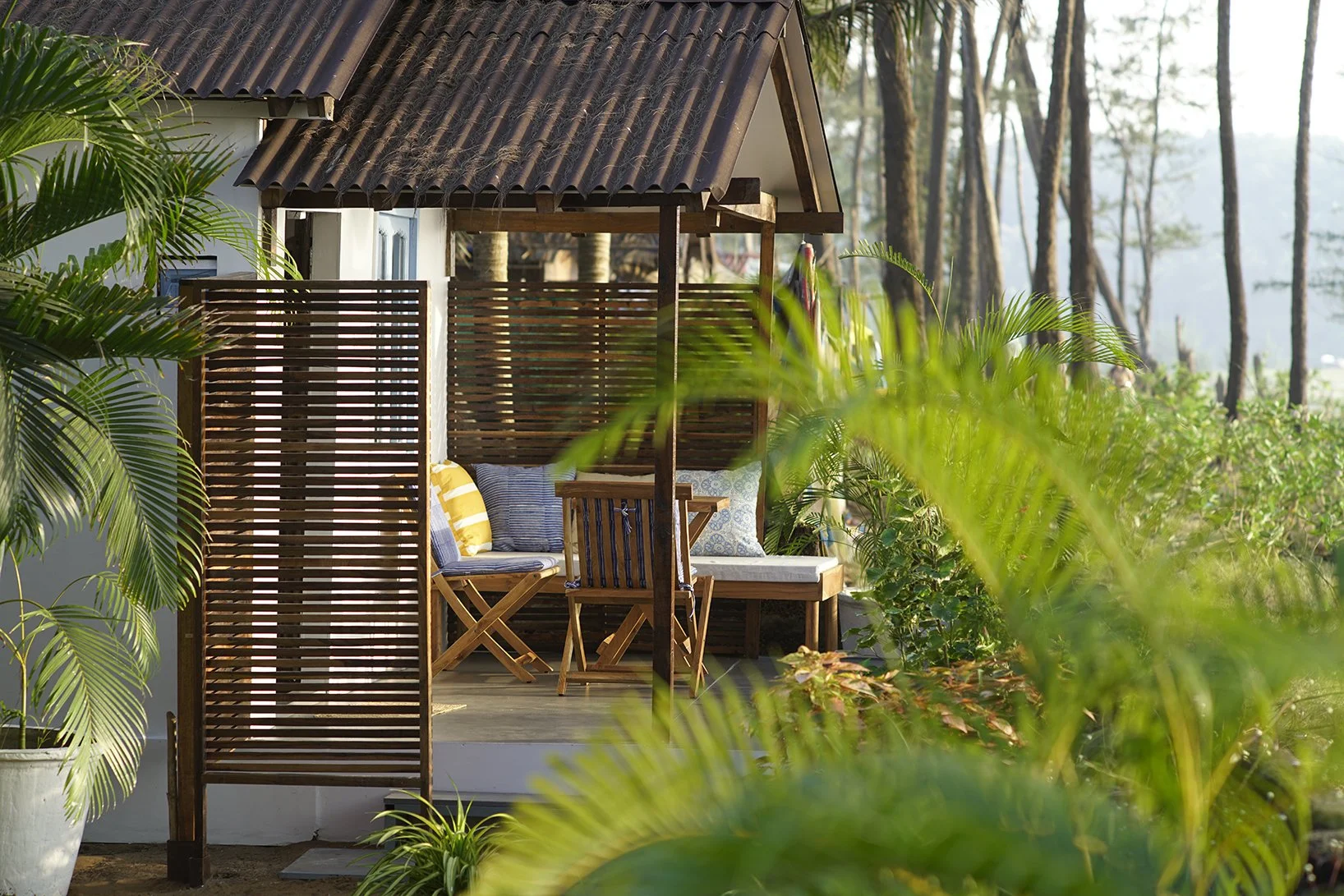 A cozy outdoor patio with wooden furniture, cushions, and potted plants in a lush tropical setting with trees in the background.