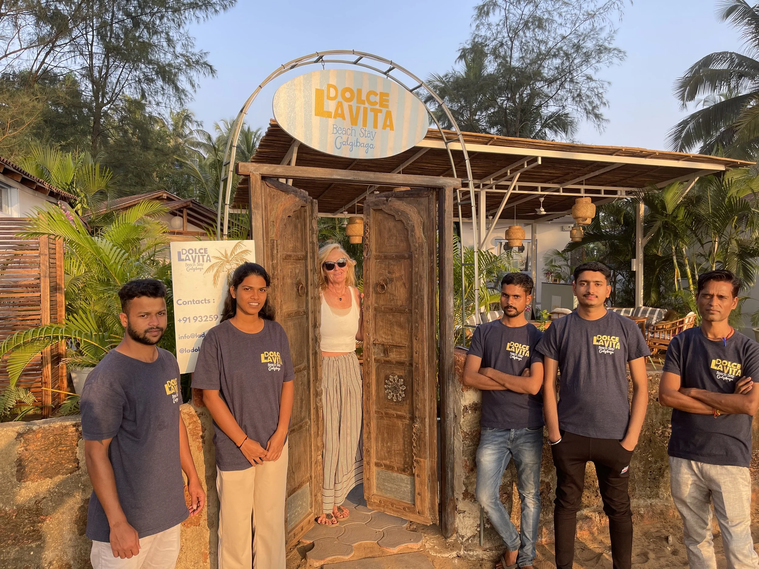 A group of six people, including five young individuals wearing matching navy blue t-shirts with the text 'DOLCE LA VITA,' standing outside a restaurant or cafe entrance named 'Dolce La Vita Beach Stay Gdalgibaga.' An older woman is standing in the doorway. The setting is tropical with palm trees and lush greenery, and the sky is clear.