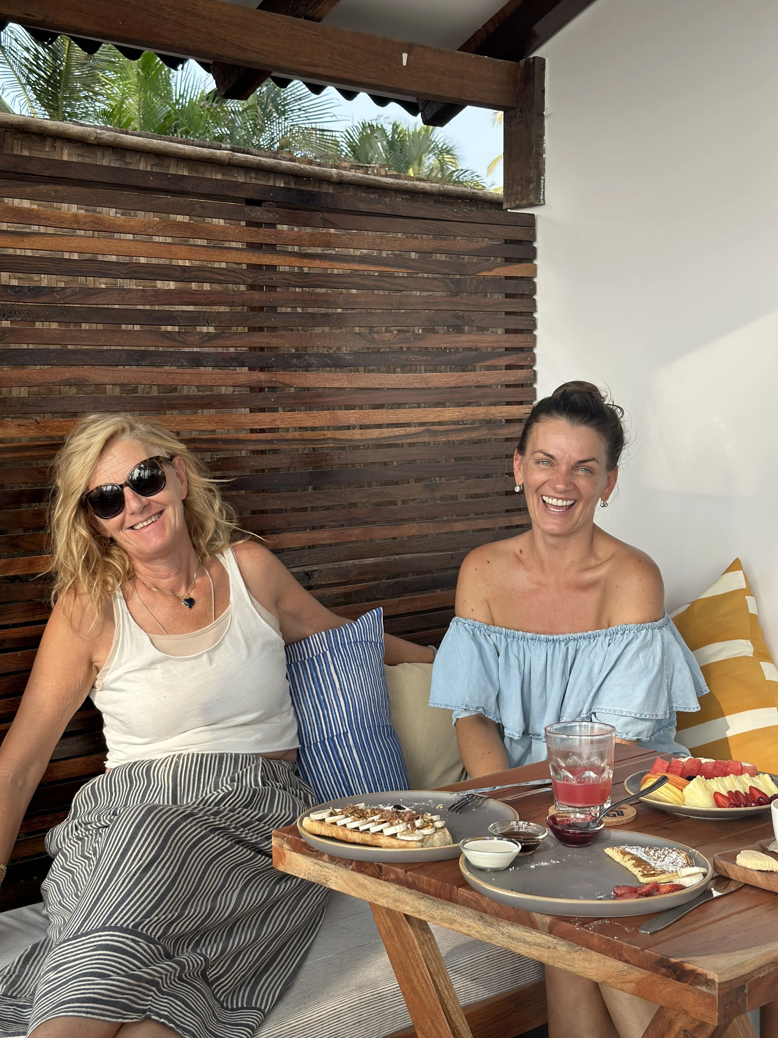 Two women sitting at a table outdoors, smiling. The woman on the left has blonde hair, wearing sunglasses, a white tank top, and a striped skirt. The woman on the right has dark hair in a bun, wearing an off-the-shoulder blue top. The table has plates of food, including fruit and desserts, and drinks.