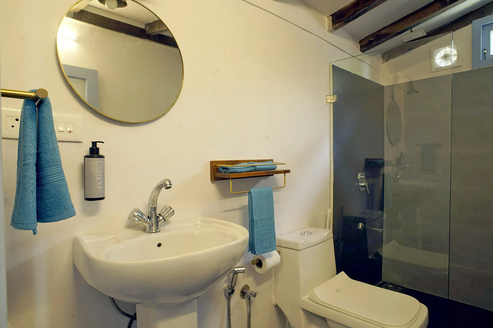 A small bathroom with a round mirror, white sink, blue towels, a soap dispenser, a toilet, and a glass-enclosed shower area with a dark wall in the background. The ceiling features exposed wooden beams.