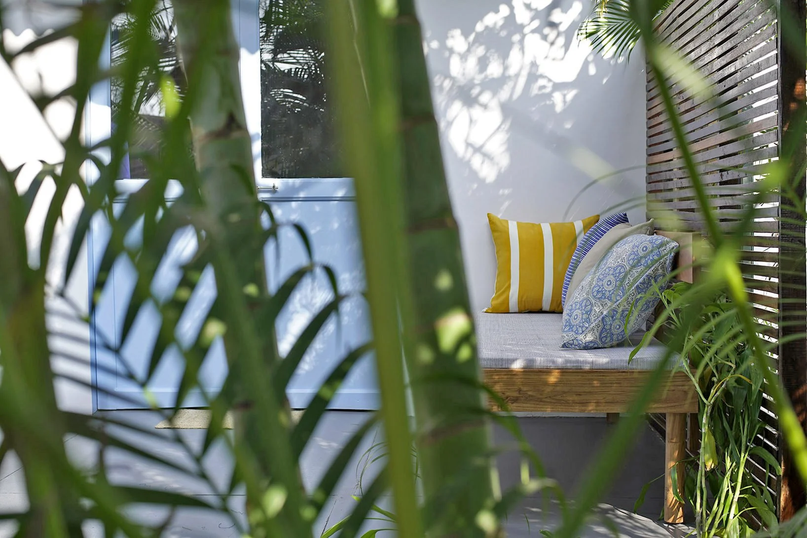 Outdoor seating area with a wooden bench, a yellow and white striped pillow,, and two patterned pillows, surrounded by green plants and partially enclosed by wooden slats.