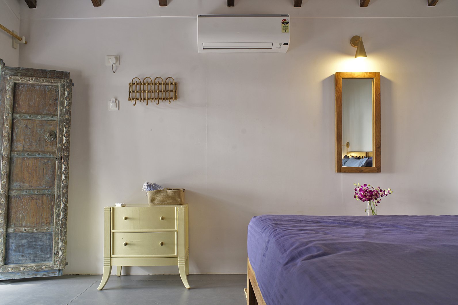 A bedroom with a bed draped in purple fabric, a small yellow nightstand, a large antique wooden wardrobe, a mirror with a wooden frame, a vase with pink flowers, a wall-mounted air conditioning unit, and a wall-mounted light. There is a decorative brass wall piece and a small white object on the nightstand.