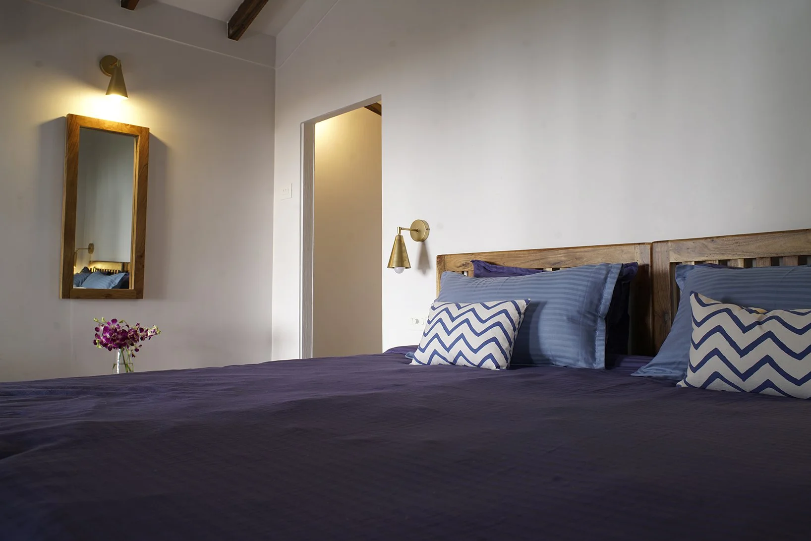A bedroom with a bed dressed in dark blue sheets and striped pillows, a wooden headboard, a small side wall lamp, a mirror framed in wood, and a vase of pink flowers on the nightstand.