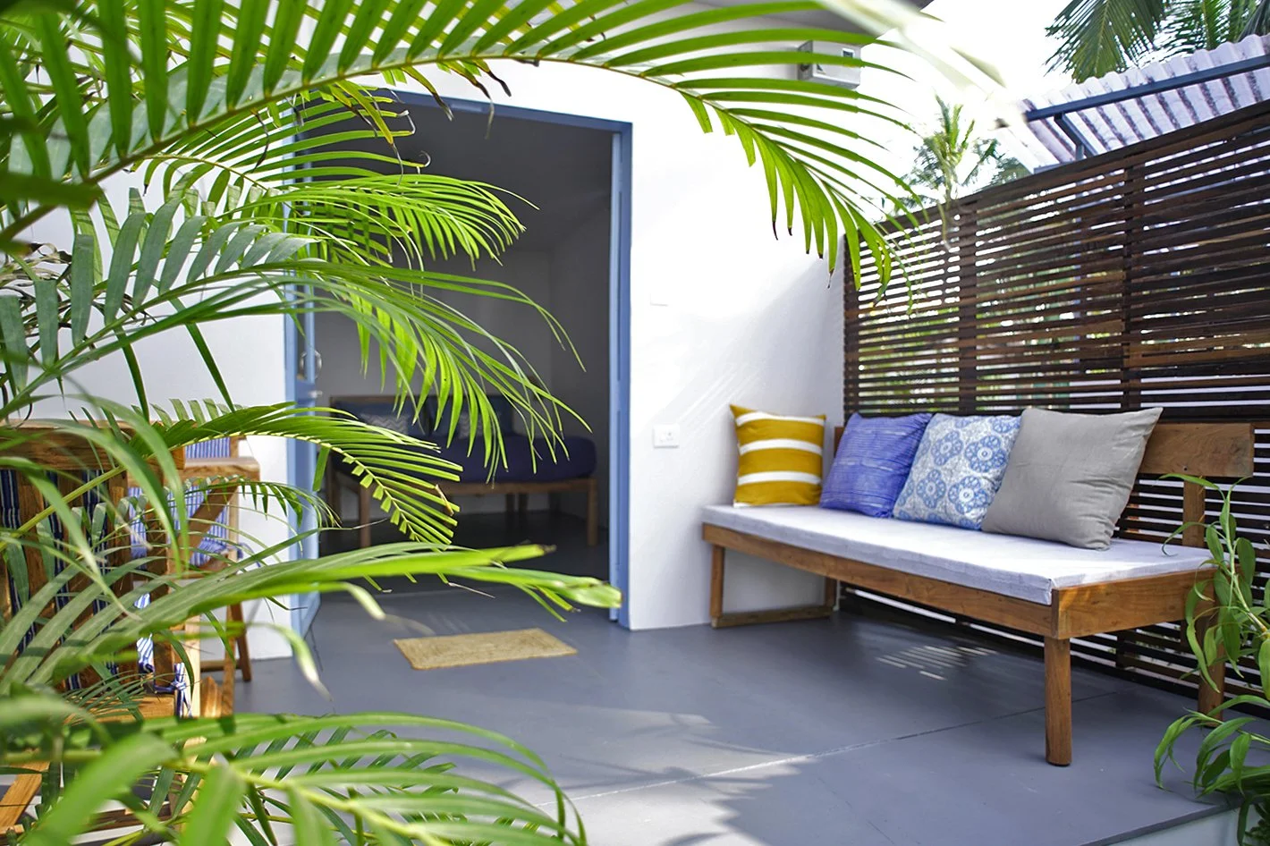 Outdoor patio with a wooden sofa, white cushions, and colorful pillows, surrounded by green plants and a wooden privacy screen.