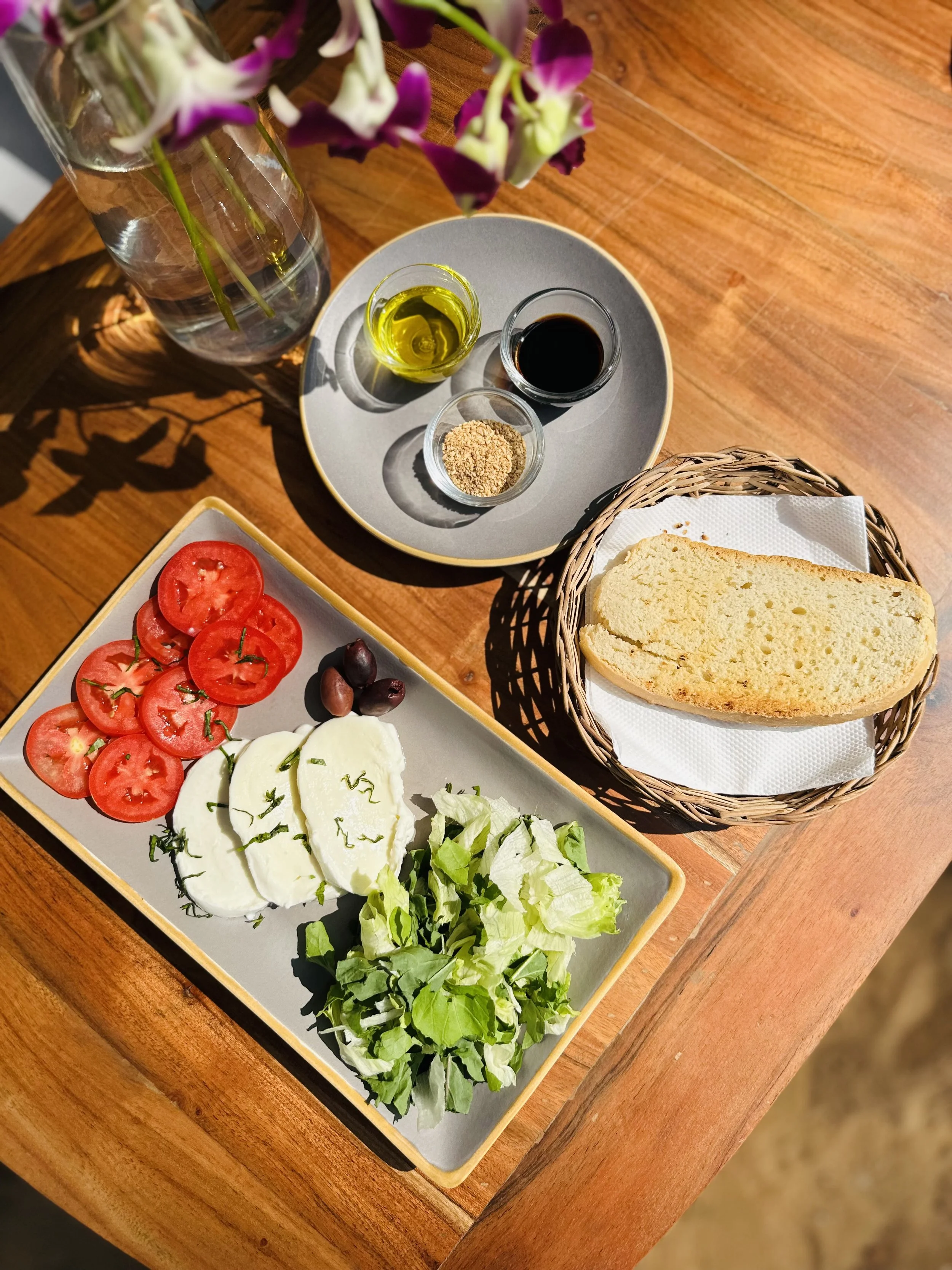 A table set with a plate of sliced tomatoes, mozzarella cheese, olives, and chopped lettuce, a basket with a slice of bread, a glass jar with pink and white flowers, and a round tray with small bowls of olive oil, balsamic vinegar, and ground seasoning.