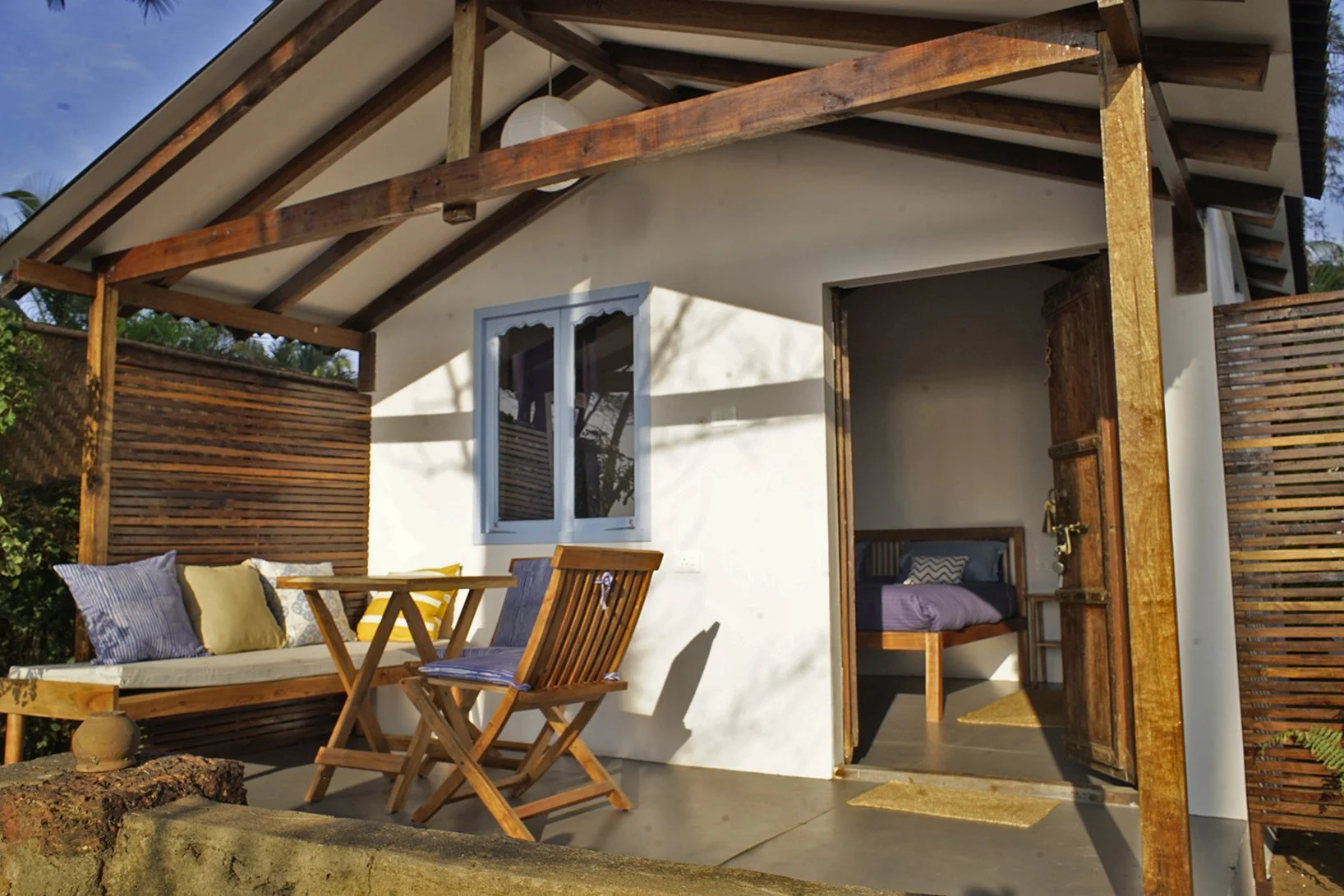 Small house with white walls and wooden accents, featuring a porch with outdoor furniture and an open door leading to a bedroom.
