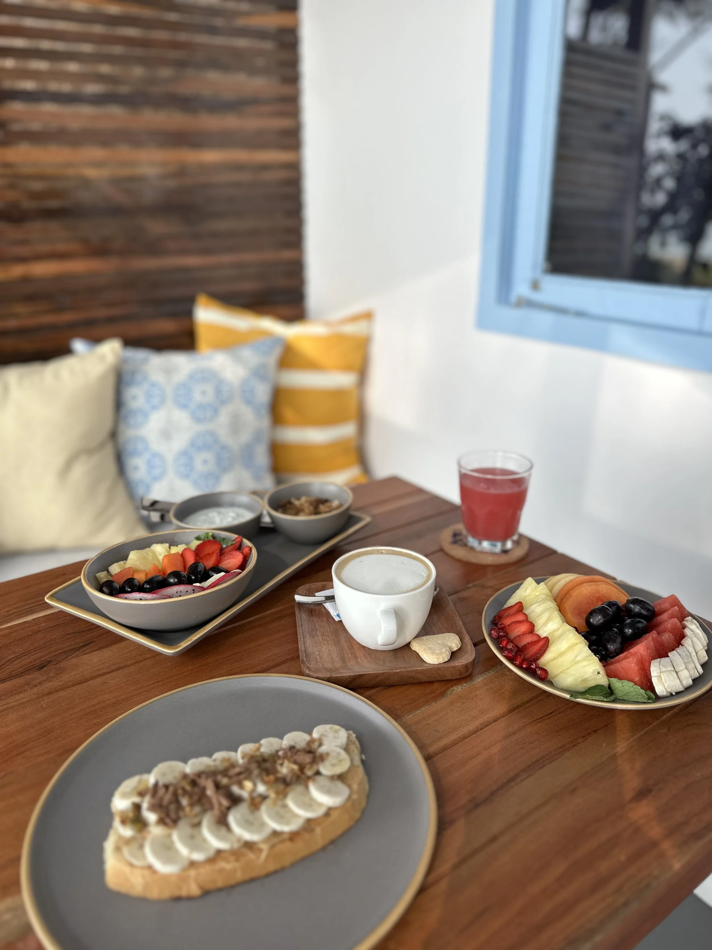 A breakfast spread on a wooden table includes a plate with a banana and nut topping, a bowl of mixed fresh fruit, a small cup of coffee, a glass of red juice, and a plate with assorted sliced fruits like pineapple, watermelon, cantaloupe, apple, and grapes, with cushions and a window in the background.