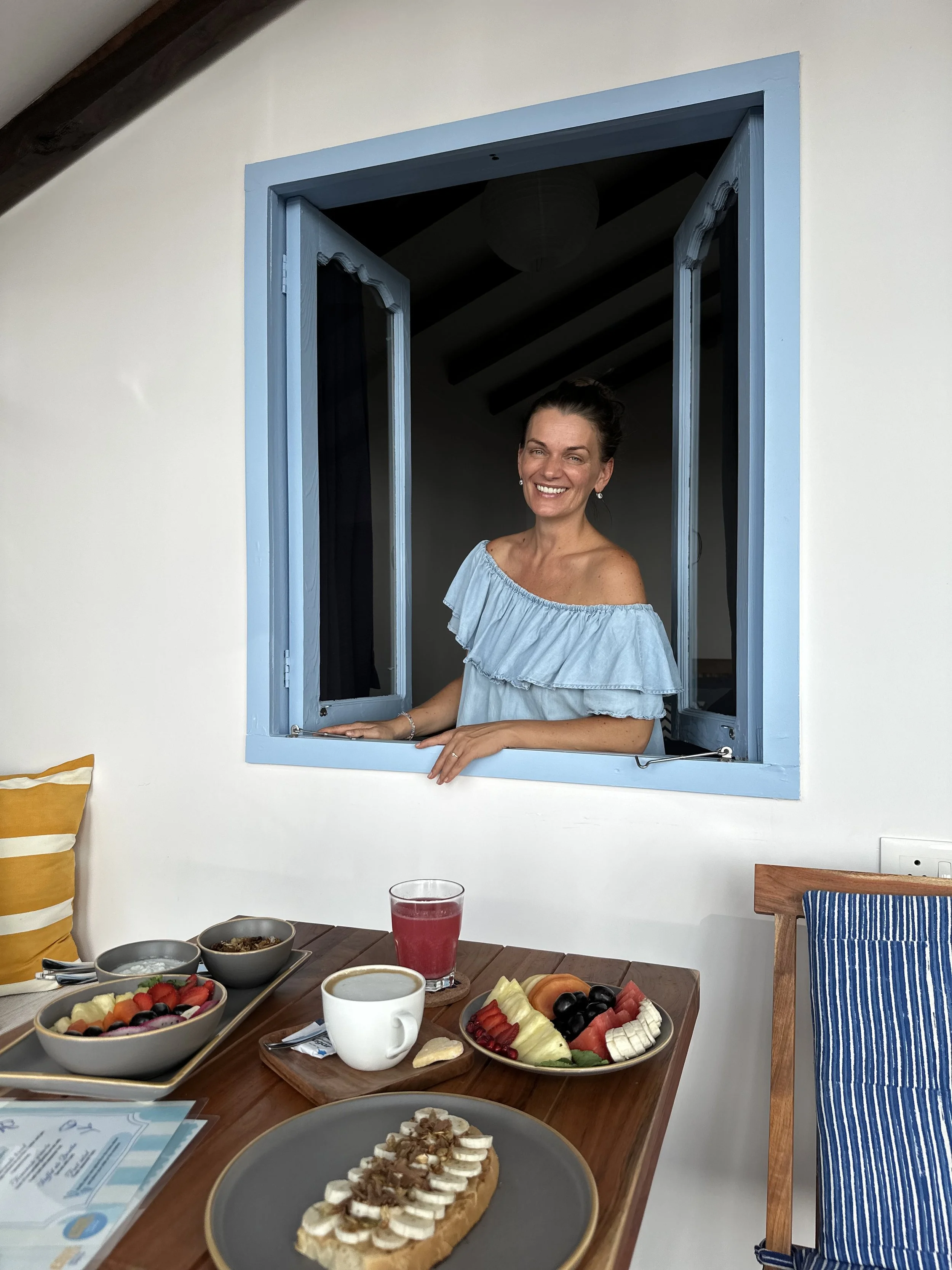 A woman is smiling and leaning out of a blue window at a breakfast table with various dishes including fruit, a croissant with toppings, and a drink.