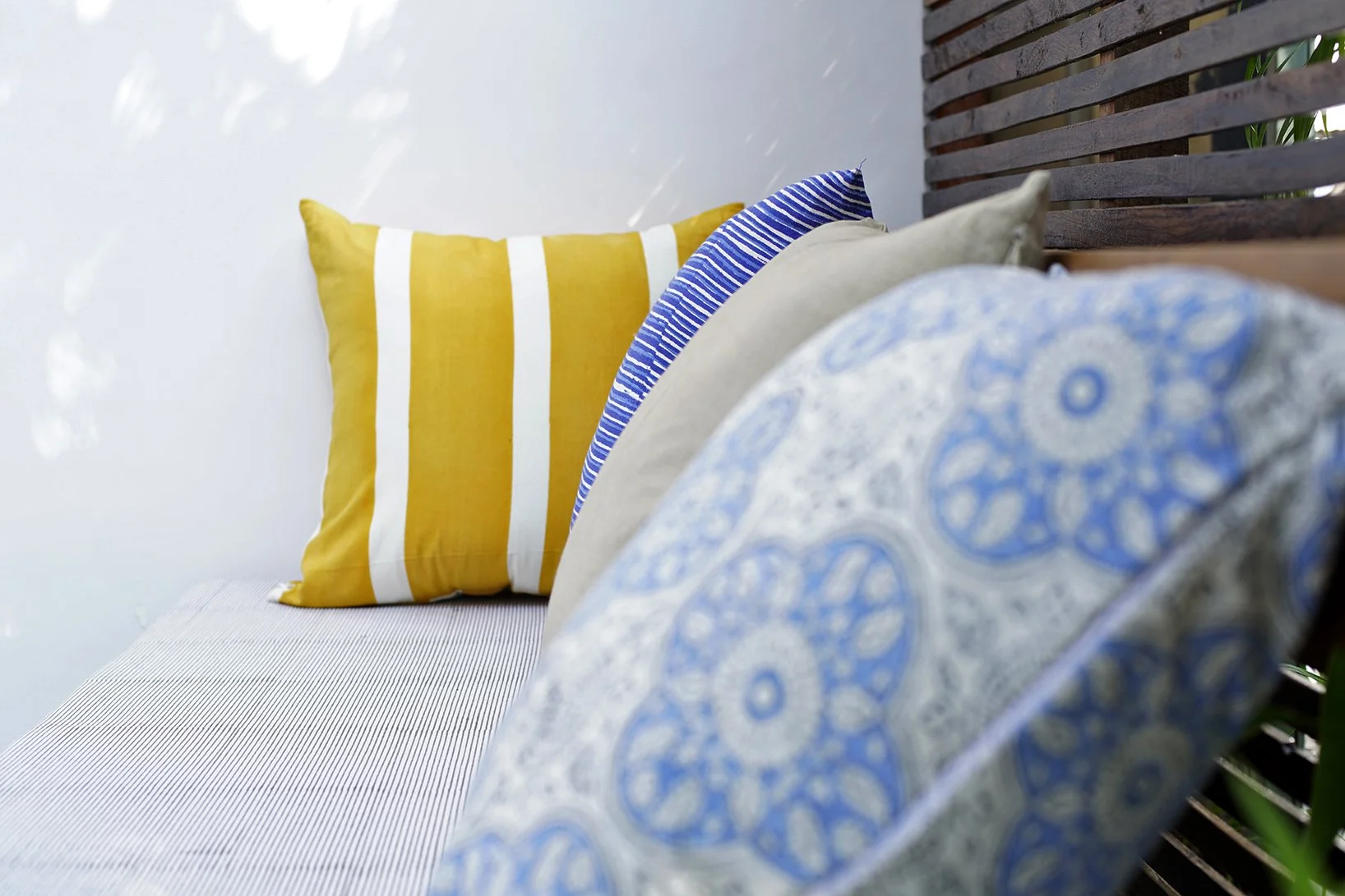 Close-up of several decorative pillows on a patio sofa, including a yellow and white striped pillow, a blue and white striped pillow, a beige pillow, and a blue patterned pillow, with a wooden slat wall and greenery in the background.