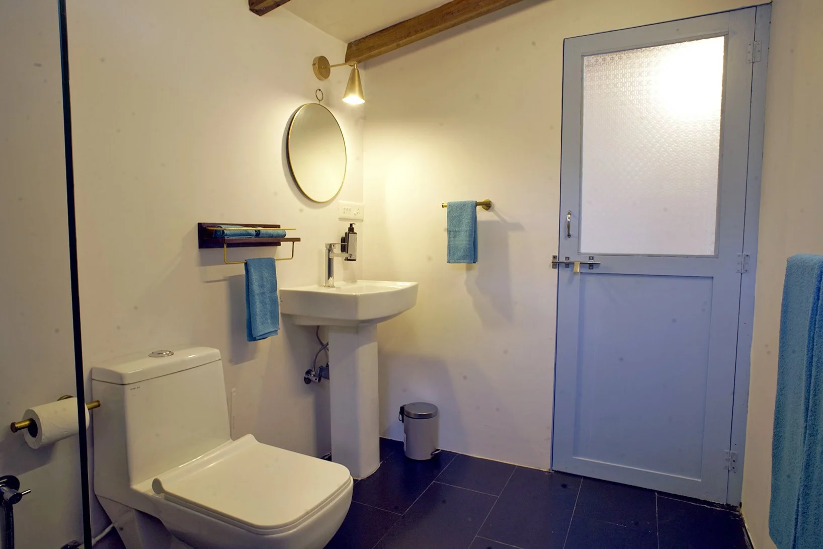 A small bathroom with white walls, a white toilet, a white pedestal sink with a mirror above, a blue towel on a rack, a small trash bin, and a blue door with a frosted window.