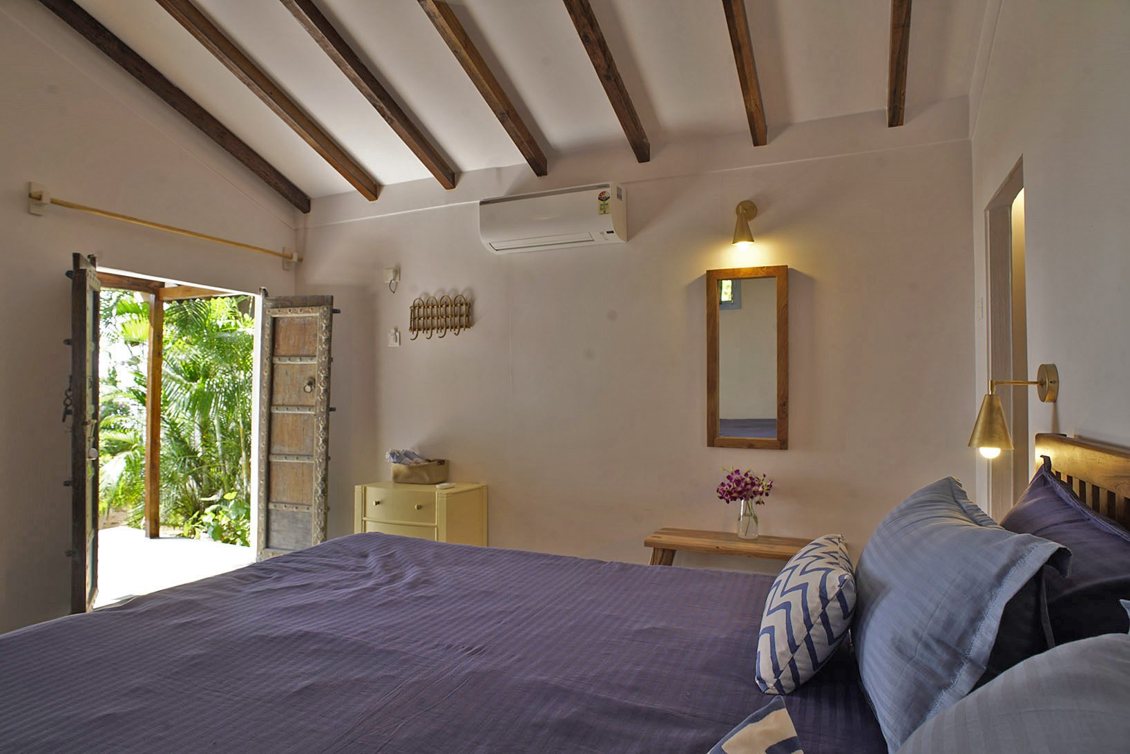 A bedroom with a large bed with purple and striped pillows. There is a door open to a balcony with greenery outside. The room has wooden beams on the ceiling, a wall-mounted mirror, a small yellow cabinet, and a small wooden table with a vase of pink