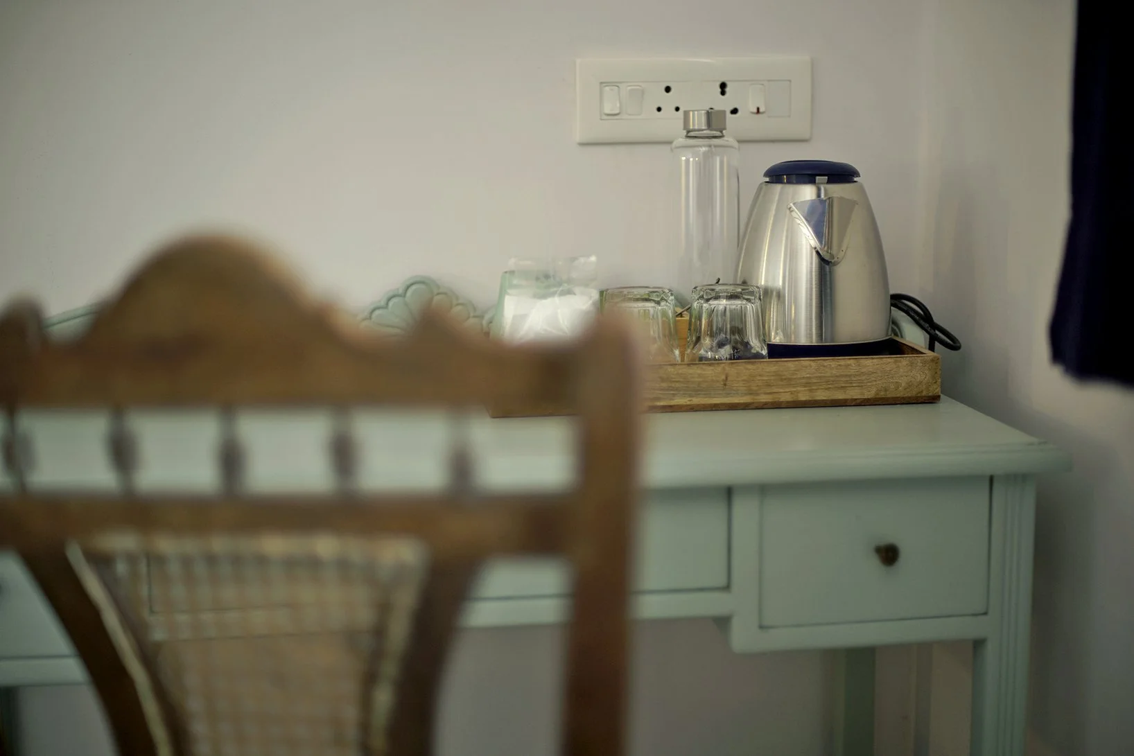 A wooden tray with a kettle, drinking glasses, and a bottle on a mint green console table in front of a white wall with a power outlet.