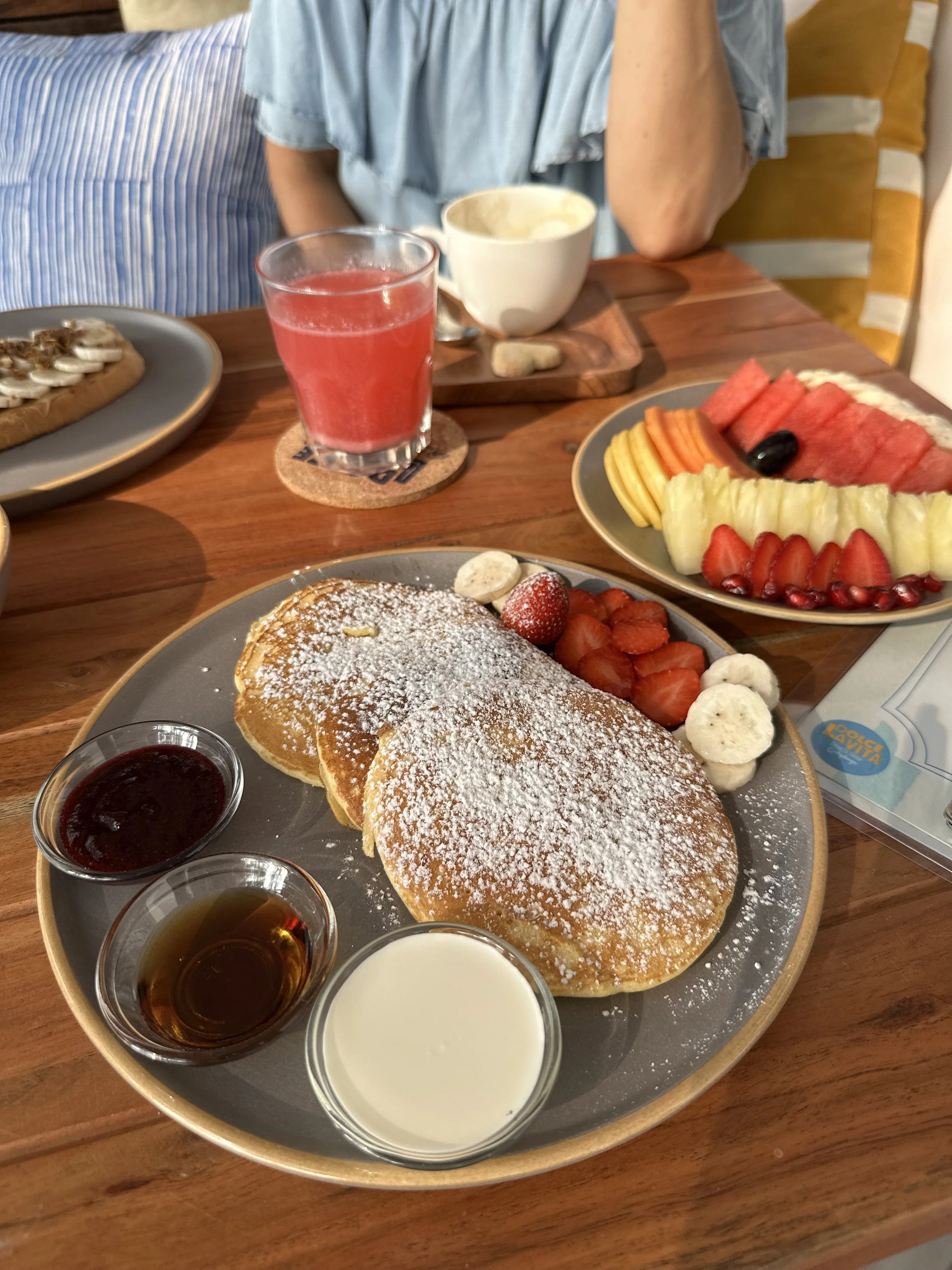 A breakfast spread on a wooden table includes a plate with three golden pancakes dusted with powdered sugar, strawberries, banana slices, and small bowls of syrup and cream. There is a plate with an assortment of sliced watermelon, honeydew, cantaloupe, strawberries, grapes, and melon balls. A glass of pink juice and a bowl of soup are also on the table, with part of a person visible in the background.