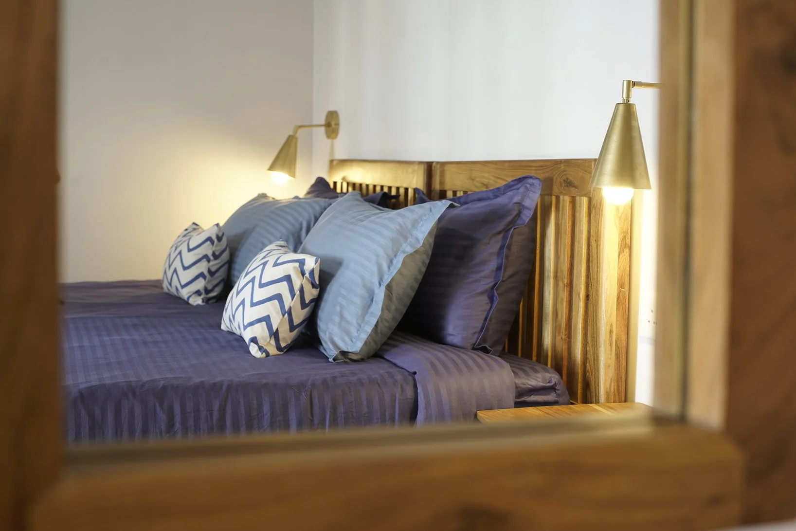 A neatly made bed with multiple pillows in different shades of blue and patterned pillowcases, captured through a mirror frame, with wall-mounted bedside lamps on both sides.
