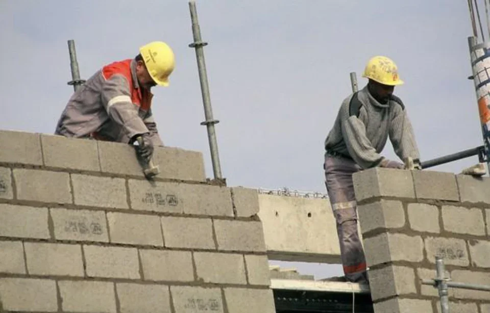 İki inşaat mühendisi veya işçi, kask ve iş elbisesi ile inşaatta duvar yaparken çalışıyor, açık gökyüzü arka planında.