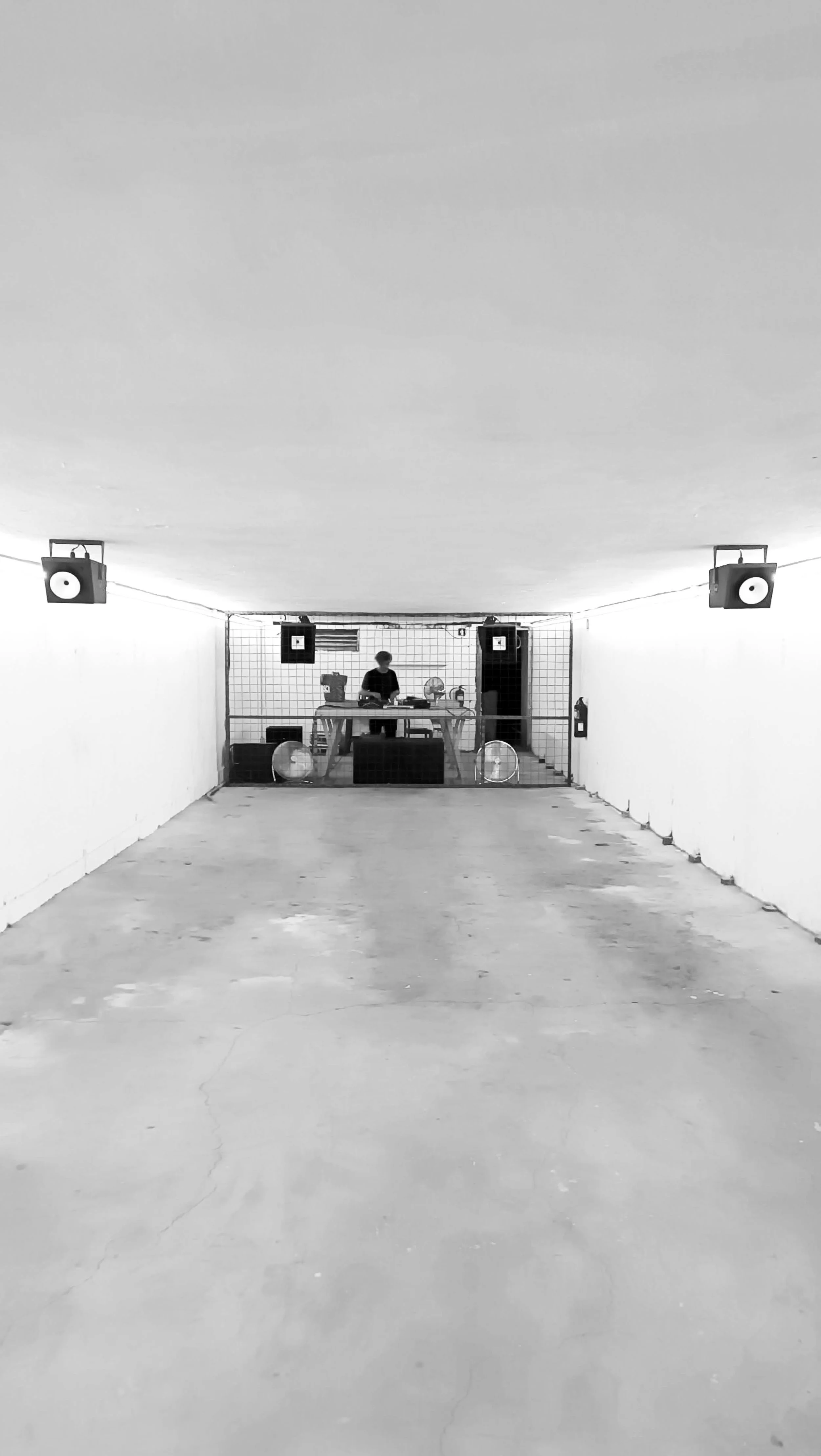 A person DJing behind a table in a dimly lit, minimalistic underground space with white walls, concrete floor, and speakers mounted on the walls.