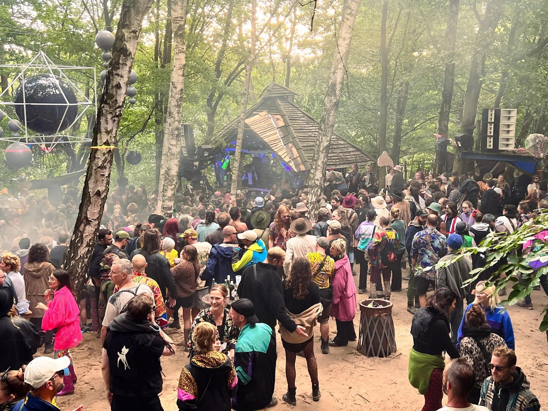 Crowd of people at an outdoor music festival in a wooded area, with a stage and DJ setup surrounded by trees.