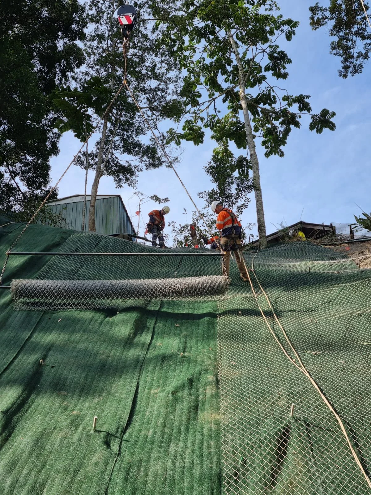 Workers wearing safety gear and high-visibility clothing are working on a construction site on a slope covered with green netting. They are secured with ropes, and one worker is near the top with a tree in the background. The sky is clear and blue.