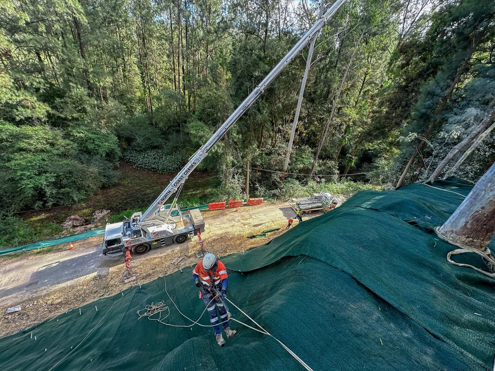 Workers installing a safety tarp on a hillside with a crane, surrounded by trees.