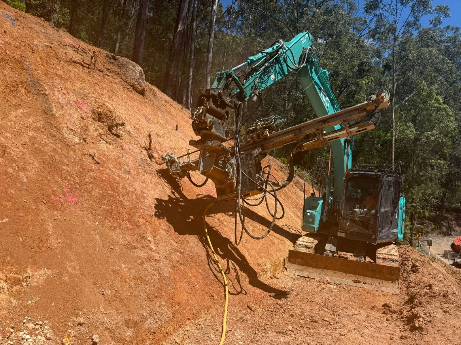 A teal-colored excavator working on a steep, reddish dirt hillside in a forested area.