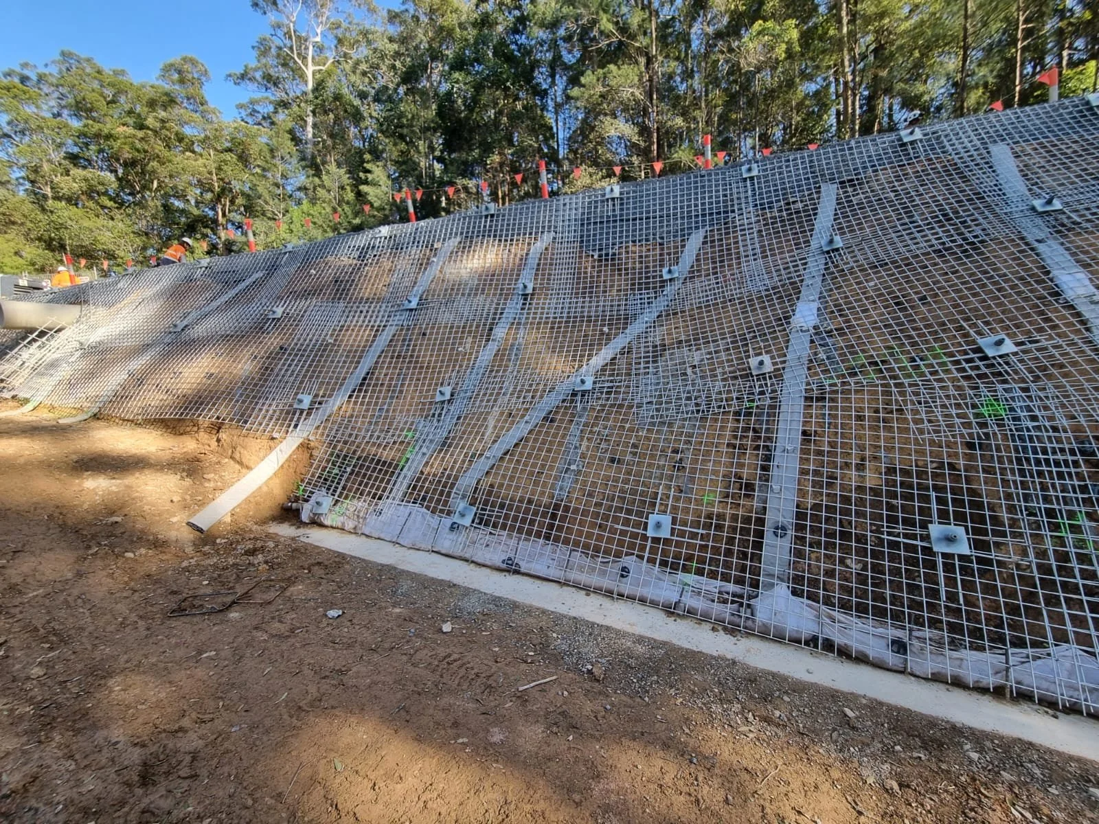 Construction site with a sloped dirt surface covered with metal grid fencing and protective netting, surrounded by tall trees and clear blue sky.
